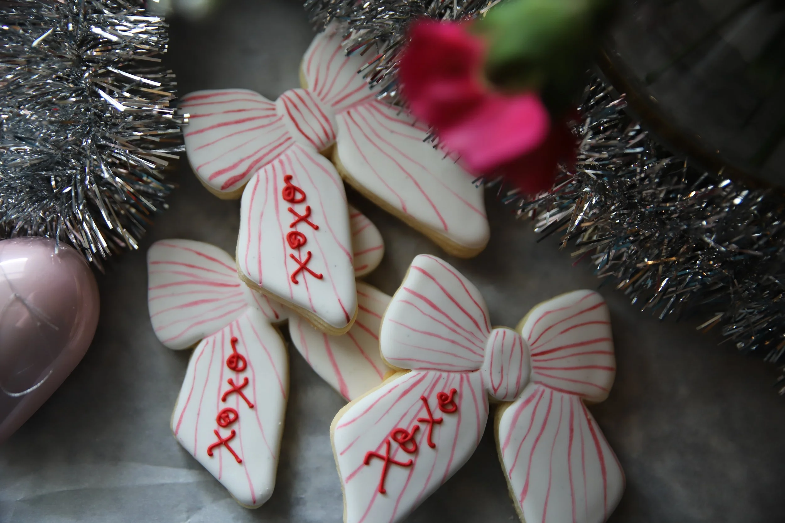 Close-up of white sugar cookies decorated with red icing in the shape of bows, surrounded by silver tinsel and a pink ornament.