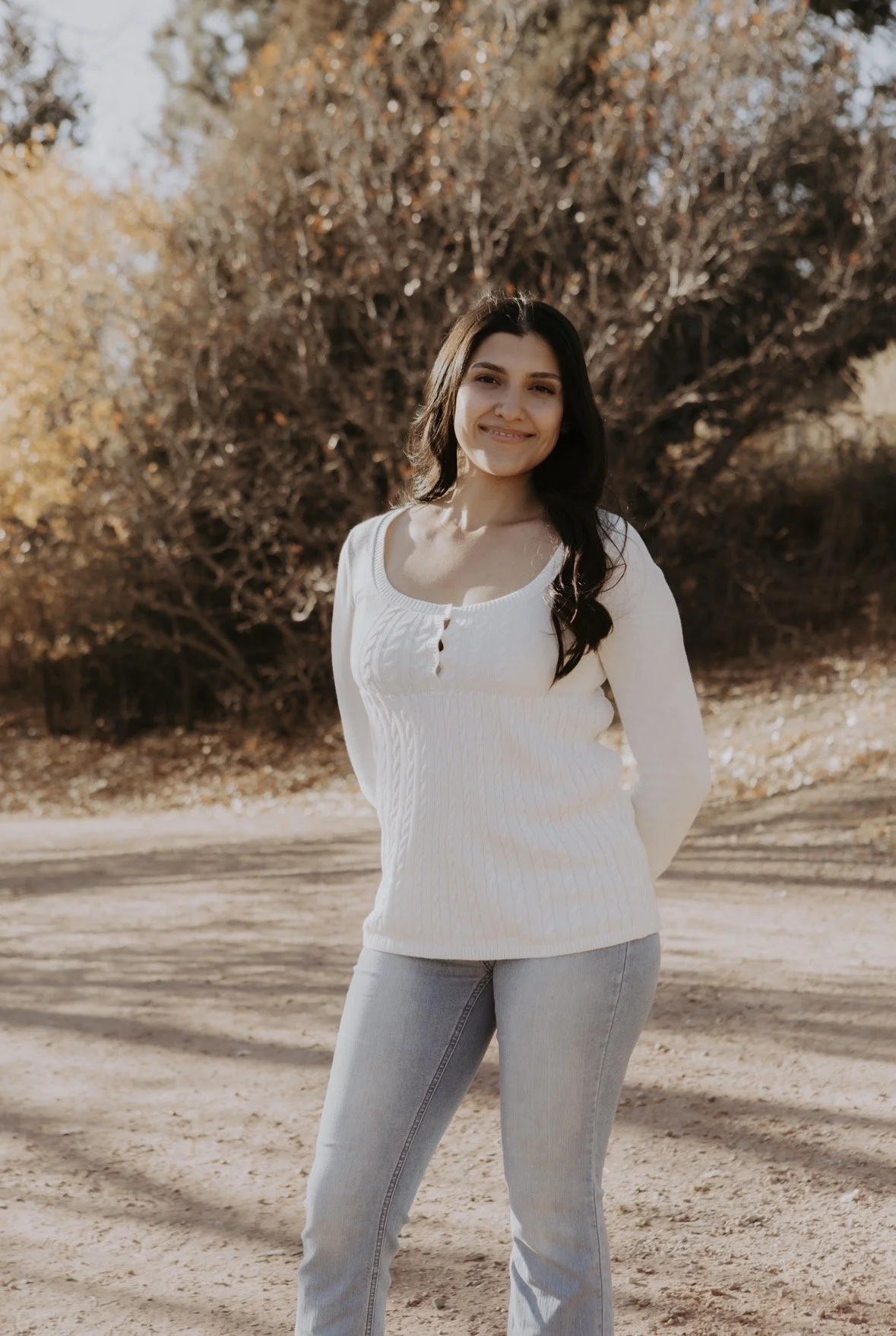 A woman standing outdoors with trees and fallen leaves in the background, smiling at the camera, wearing a white sweater and light gray jeans.