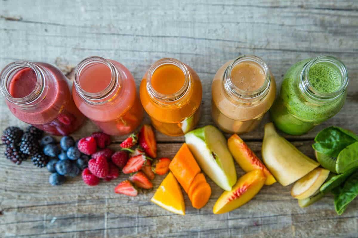 Rainbow assortment of fresh fruit and vegetable juices with berries, citrus, melon, carrots, peaches, and leafy greens arranged on a rustic wooden table.