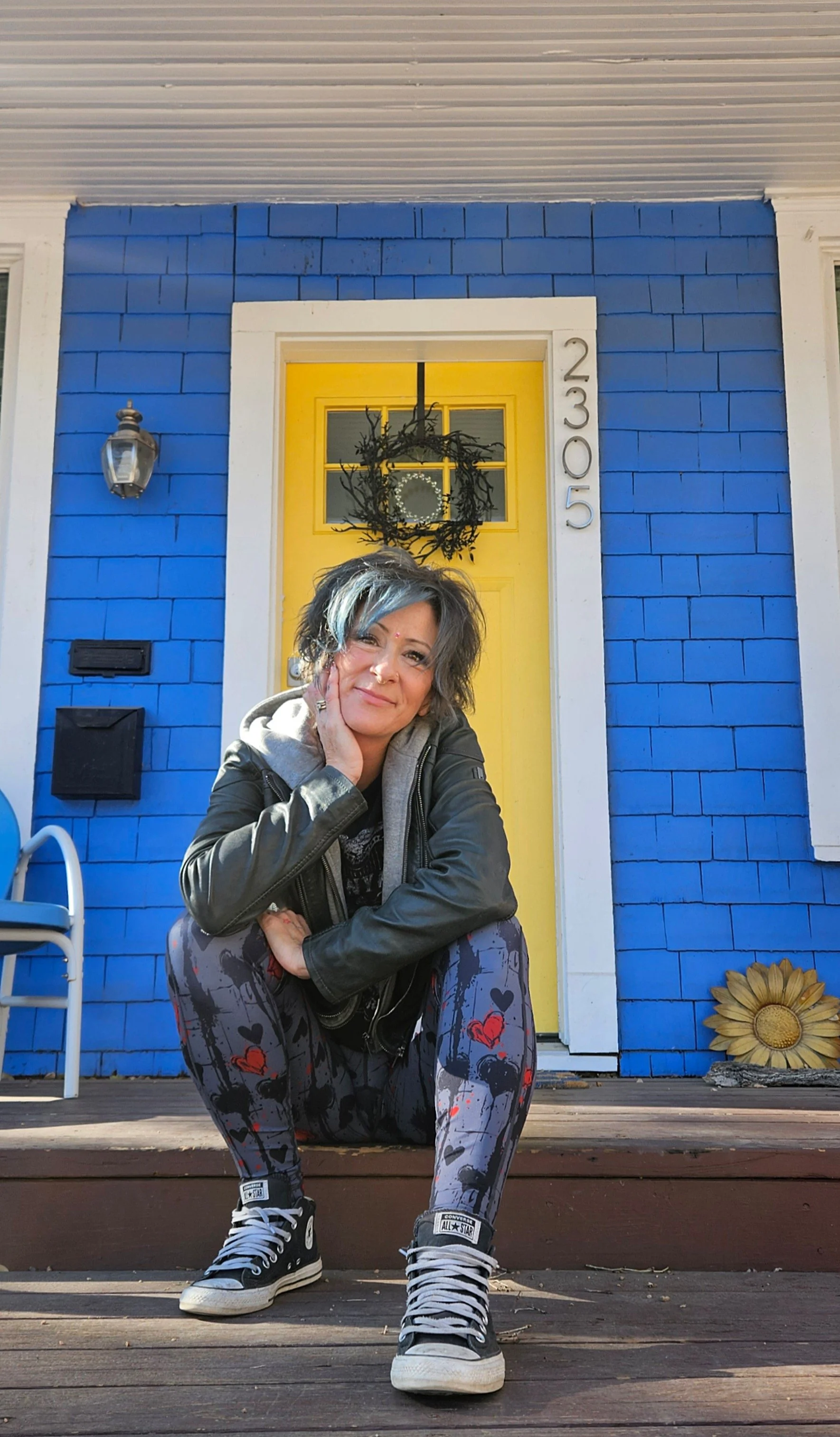 Woman with gray and blue hair, wearing a gray hoodie, black leather jacket, patterned leggings with hearts, and black Converse sneakers, sitting on a porch in front of a yellow door with a wreath, blue exterior wall, and house number 2305.