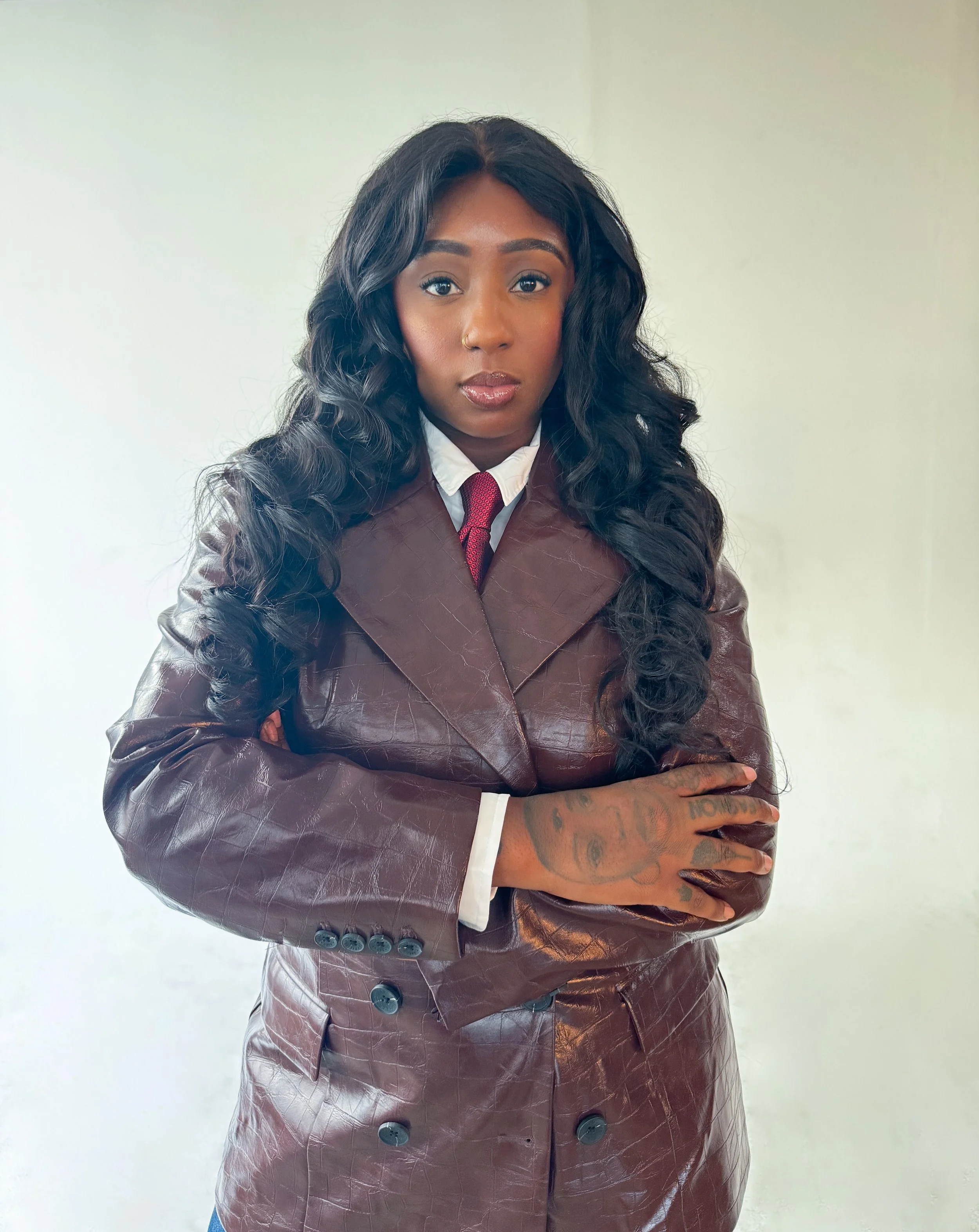 A woman with long, curly black hair, wearing a brown leather coat with a white shirt and a red tie, standing against a plain white background.