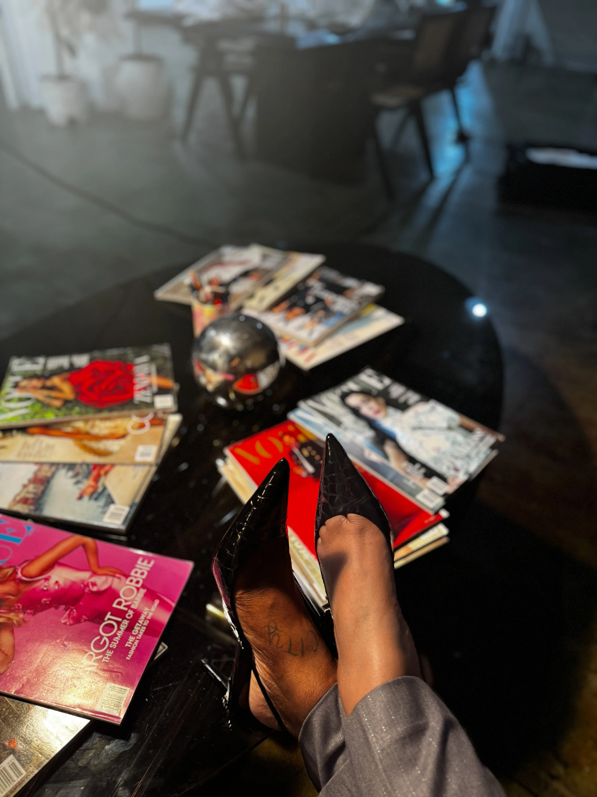 Person holding a black and pink BDSM paddle in front of a table with magazines, in a room with a polished concrete floor and chairs in the background.