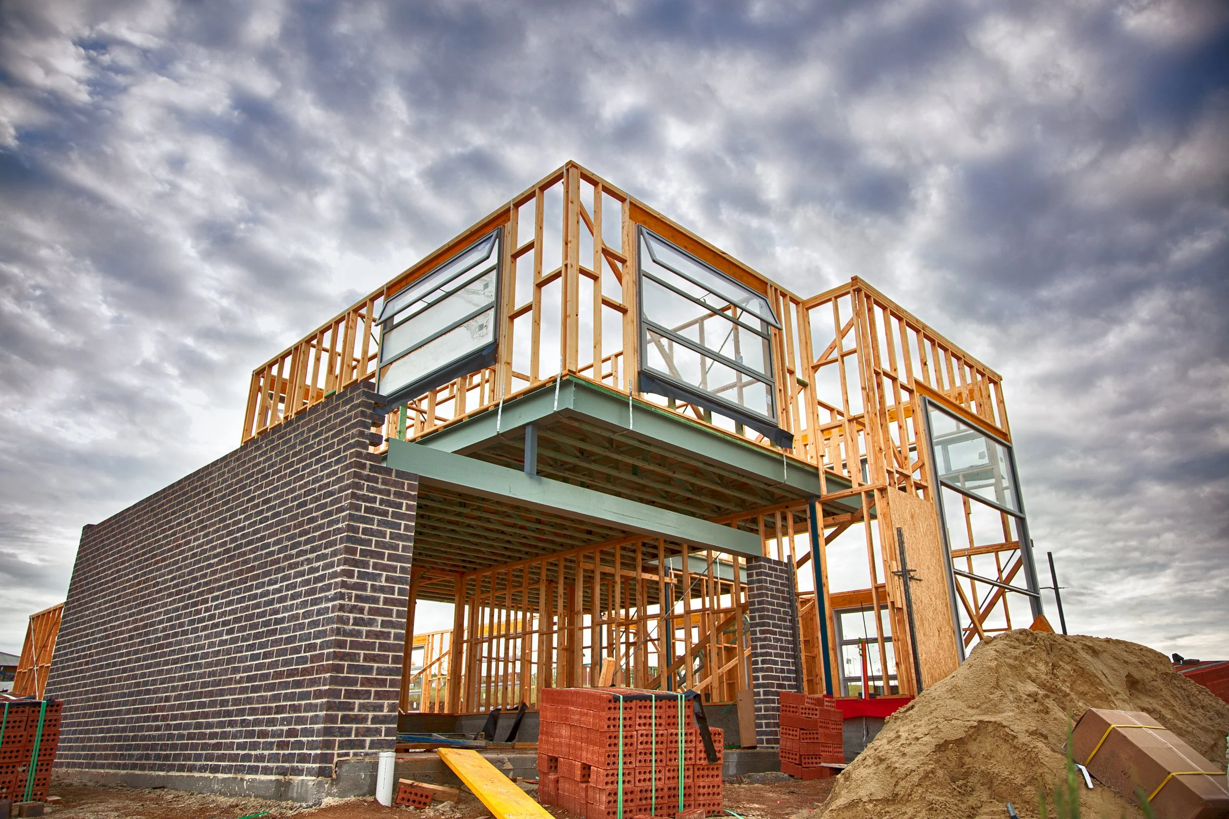 Construction site of a modern building with brick exterior walls partially built, wooden framing, and large windows, under cloudy sky.