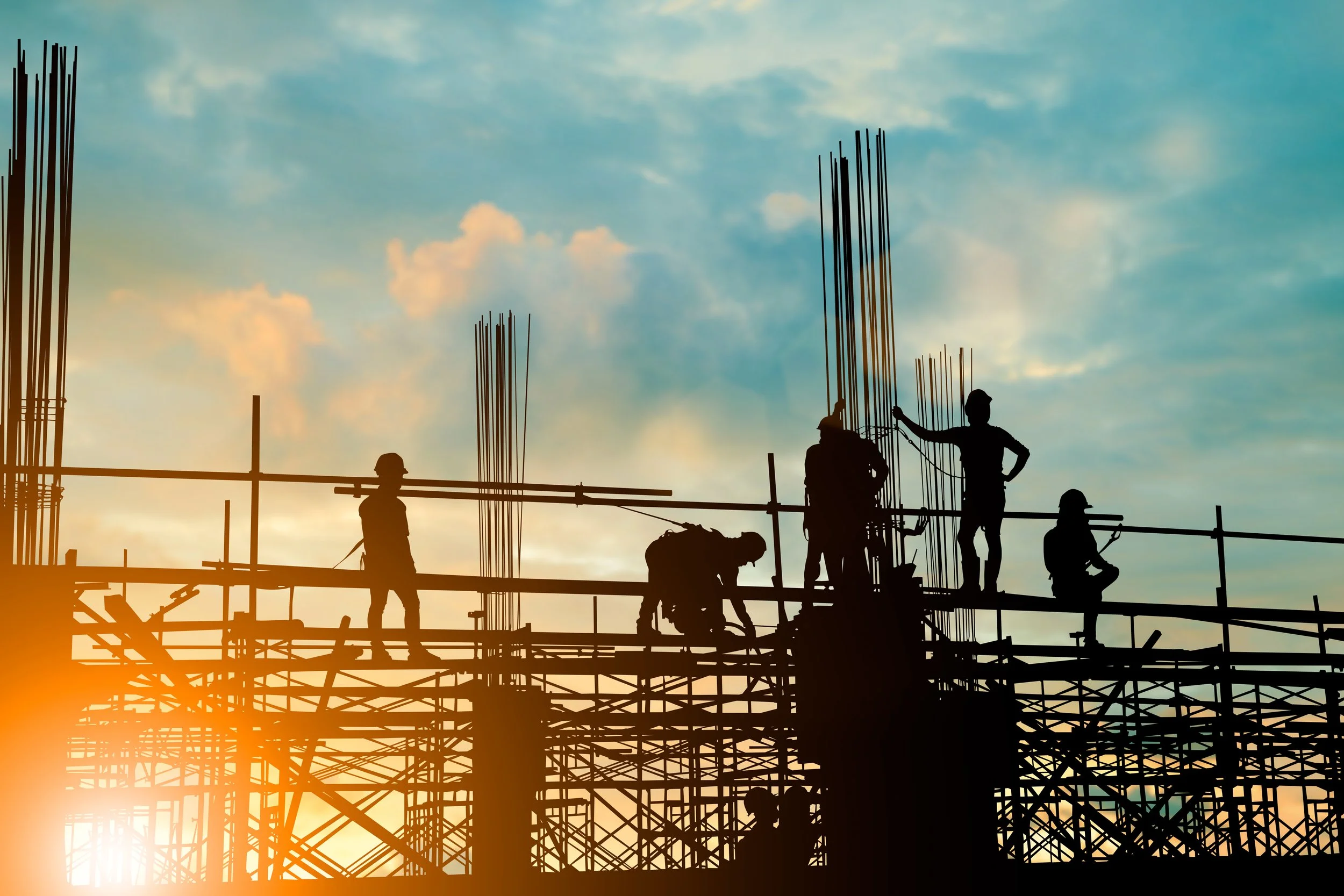 Silhouettes of construction workers working on a building construction site during sunset, with reinforced steel bars protruding from the structure.