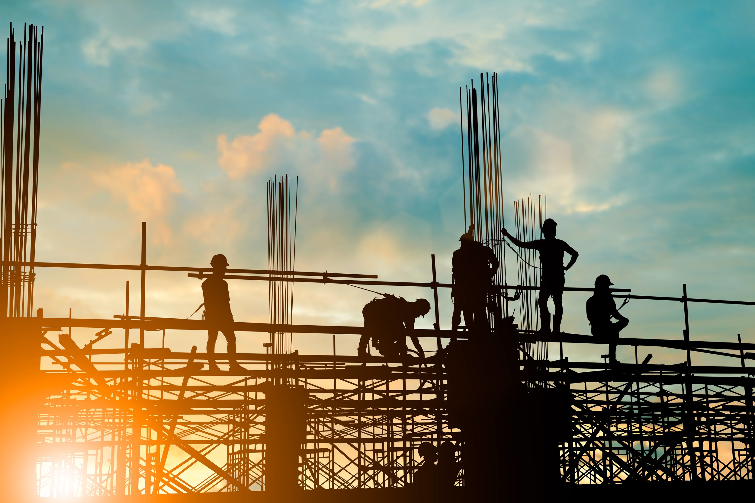 Silhouettes of construction workers on scaffolding during sunset, with steel rebar protruding from the structure and a partly cloudy sky in the background.