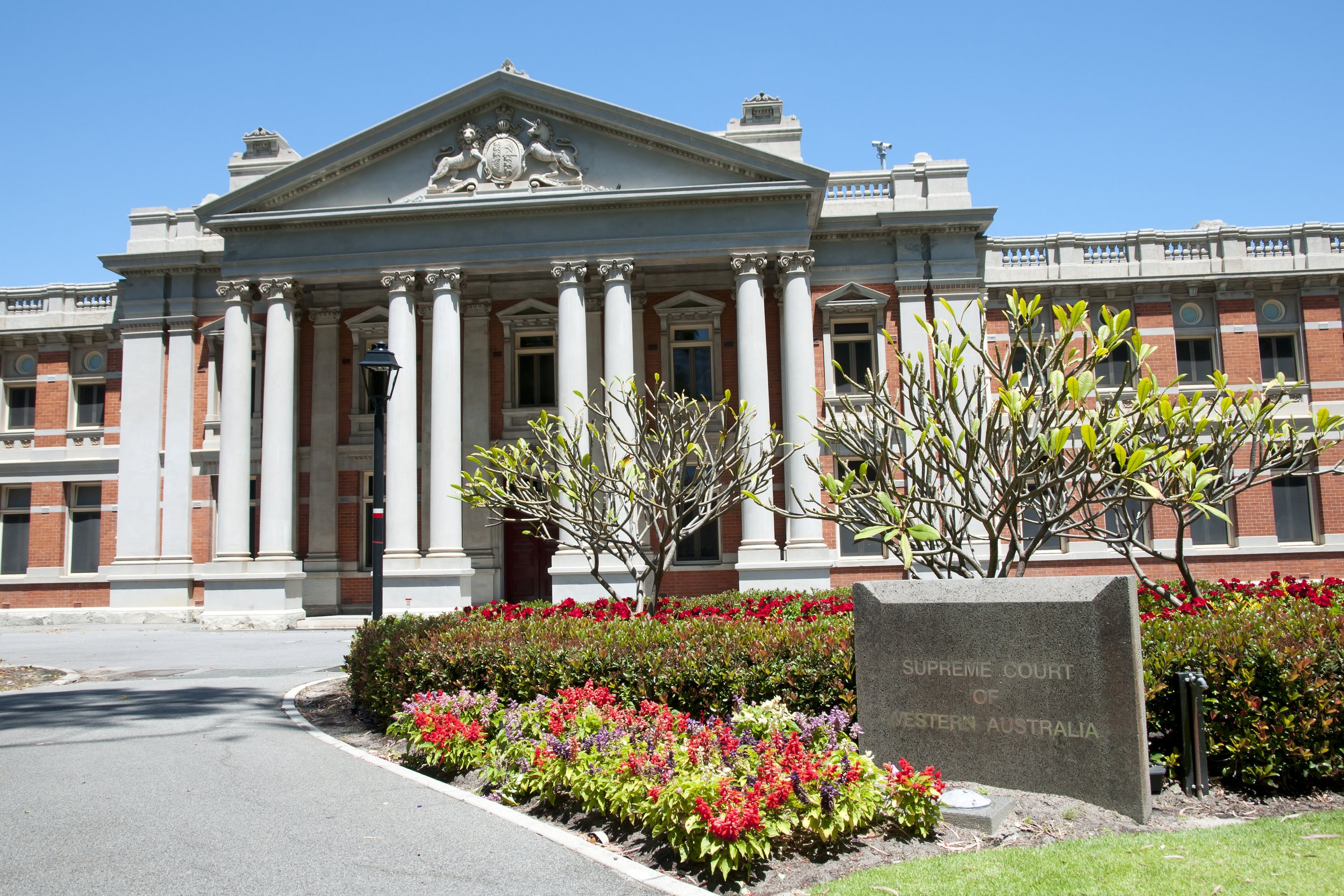 Historic building with white columns and a triangular pediment showing a crest with lions, surrounded by red brick walls, with a foreground of colorful flowers and shrubbery, and a stone sign that reads "Supreme Court of Western Australia" under a clear blue sky.