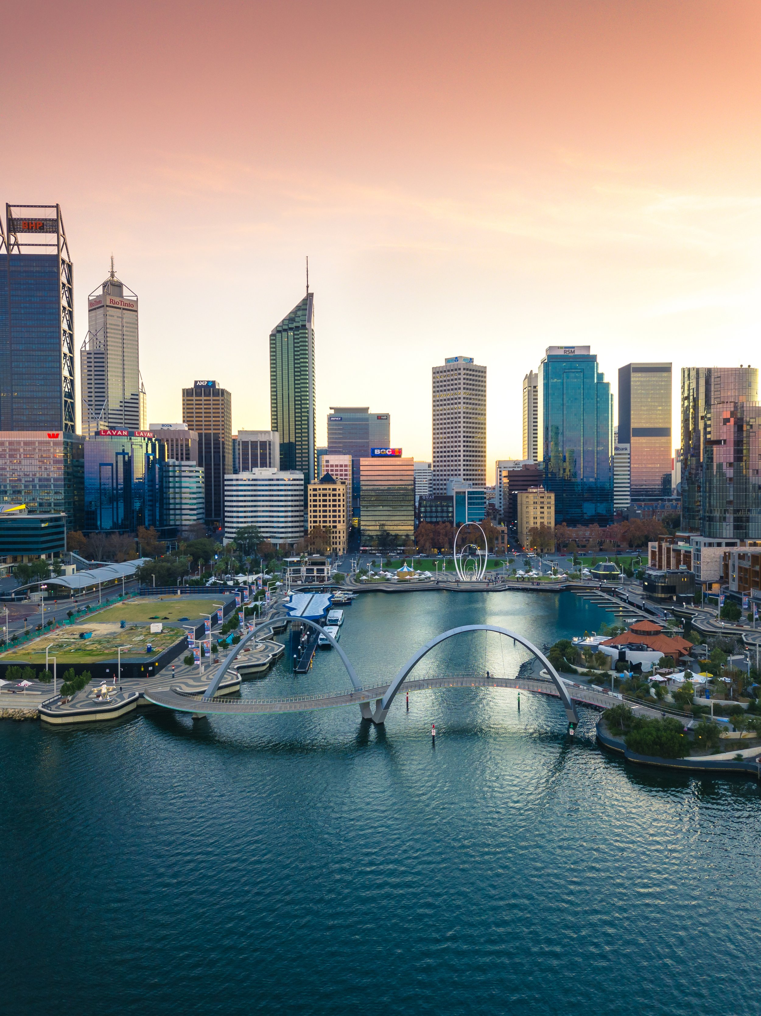 A city skyline at sunset with tall skyscrapers, a water body in the foreground, and modern architectural structures including a bridge over the water.