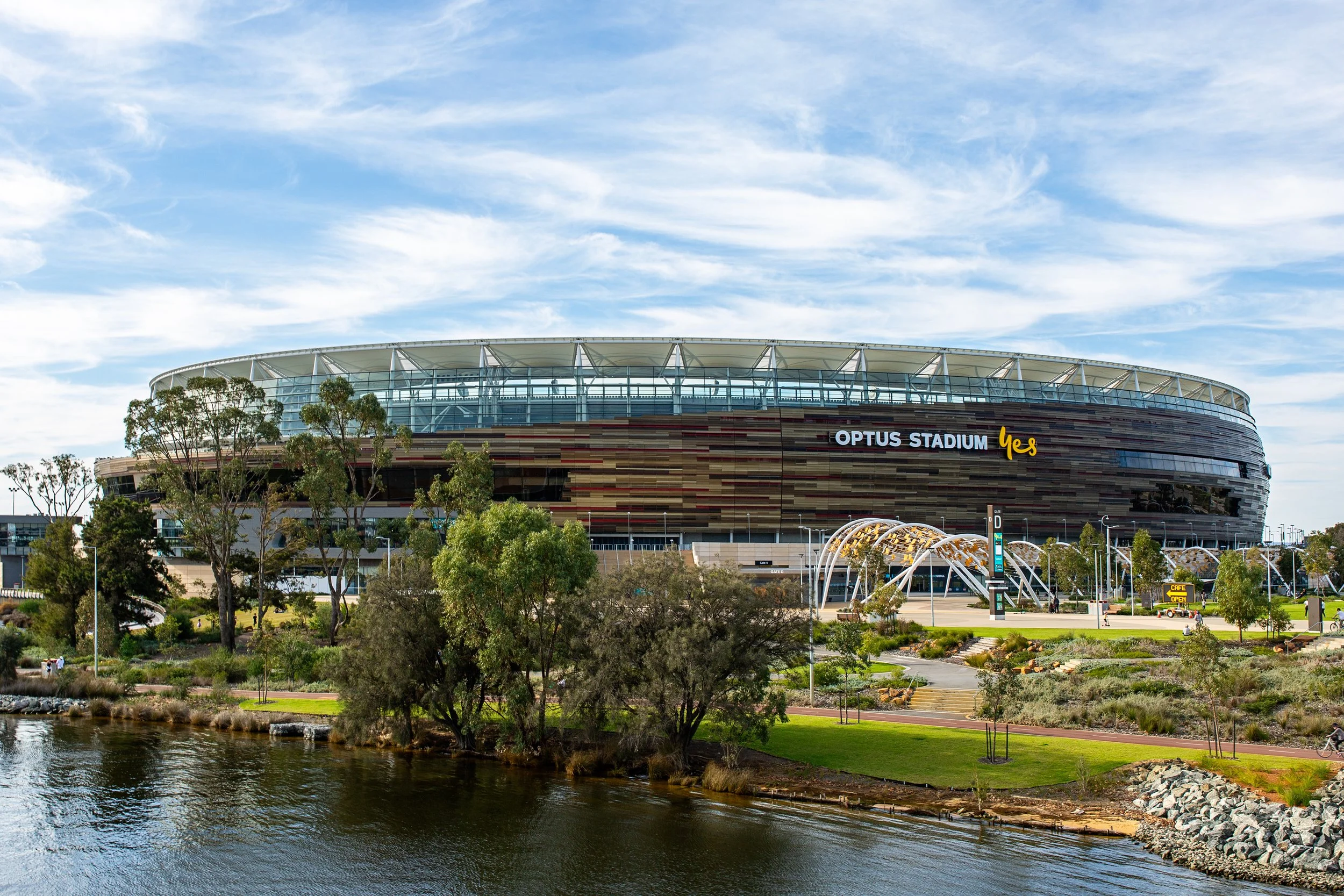 Optus Stadium in Perth, Australia, viewed across a body of water with trees in the foreground and a partly cloudy blue sky.