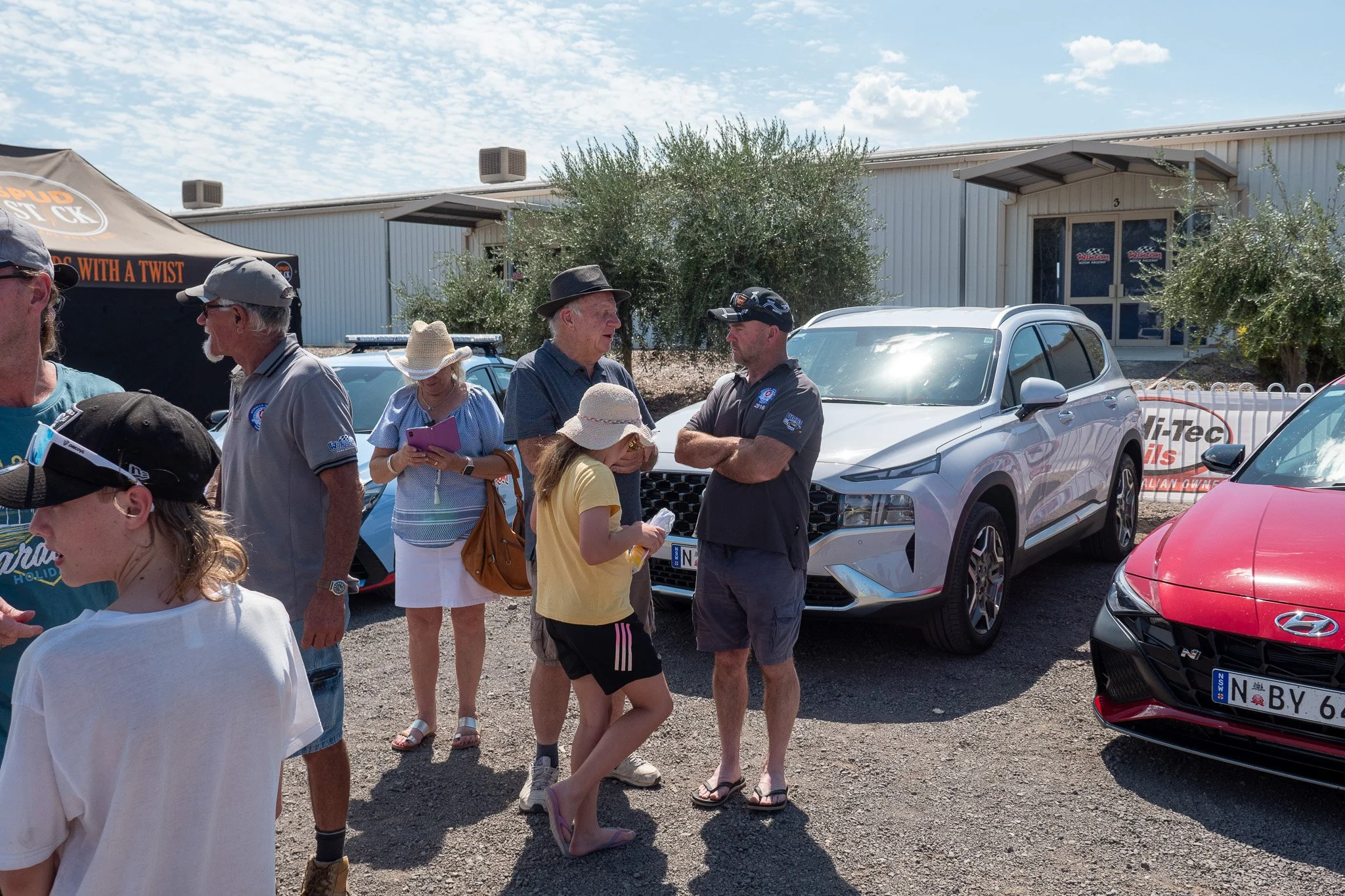 Group of people standing and talking outside near parked cars, including a white SUV, under a partly cloudy sky.