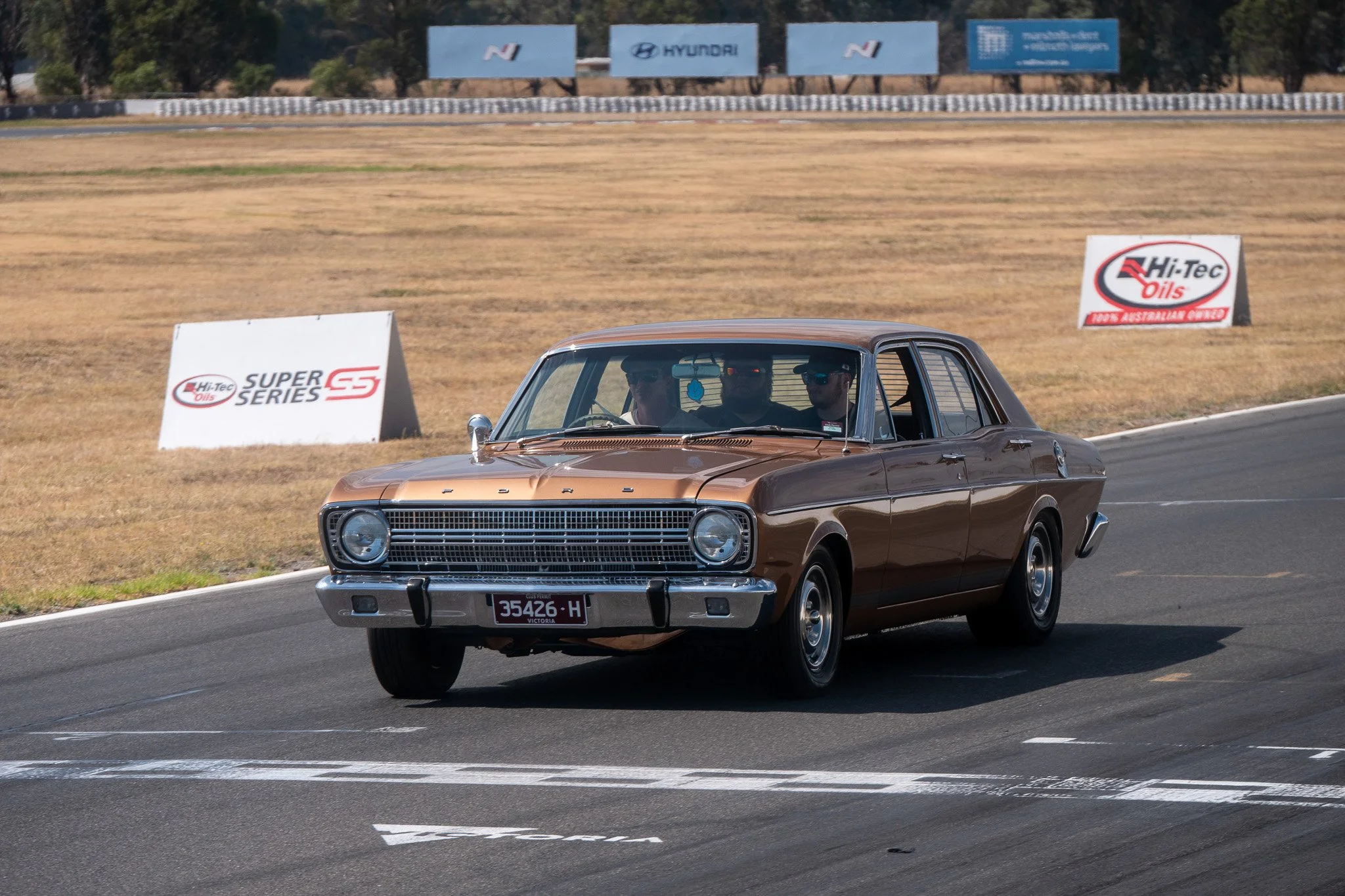 A vintage brown and chrome Ford car driving on a race track with three people inside, wearing sunglasses. The background shows advertising banners and a dry grassy field.