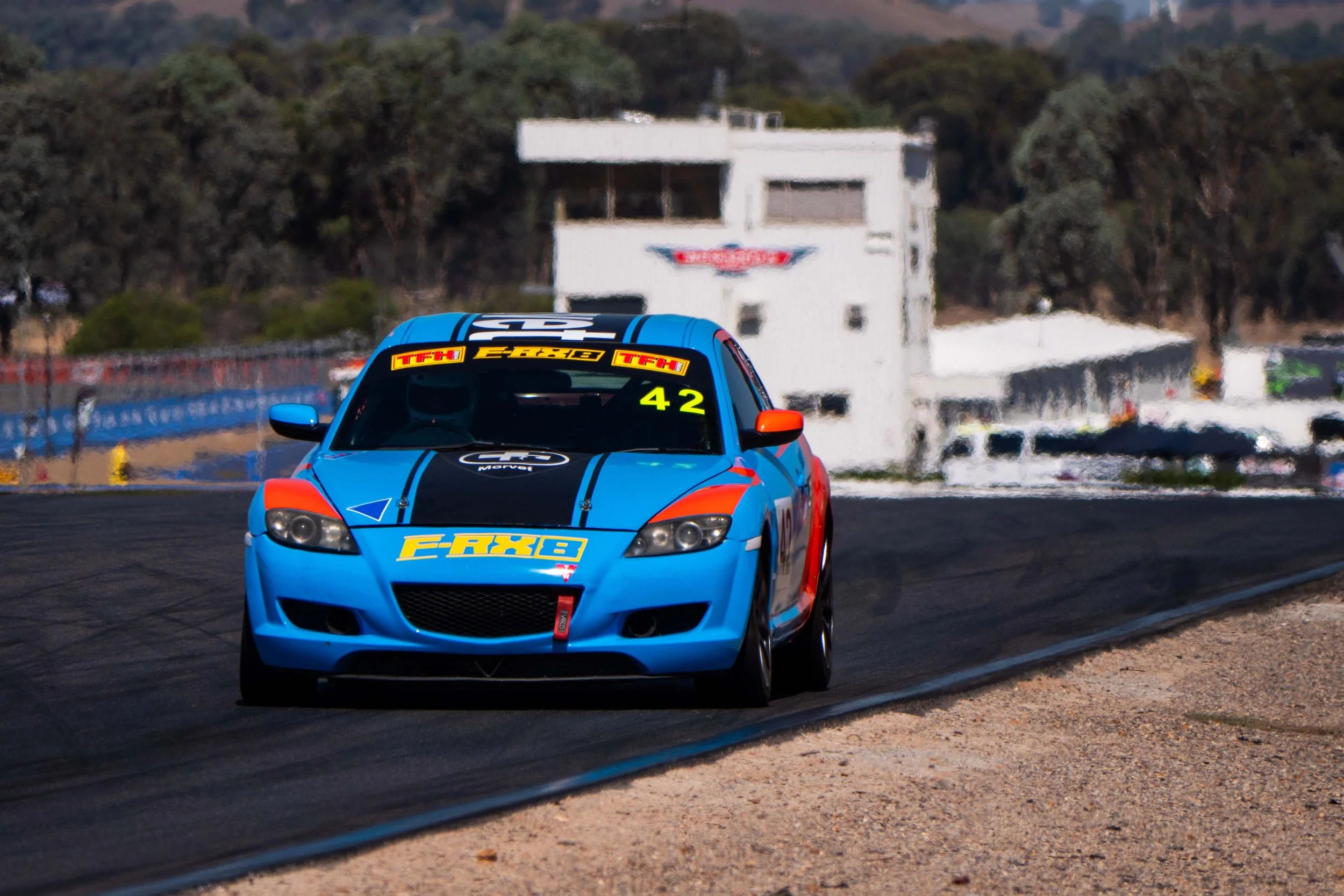 A blue race car with orange side mirrors and black and yellow decals driving on a race track.