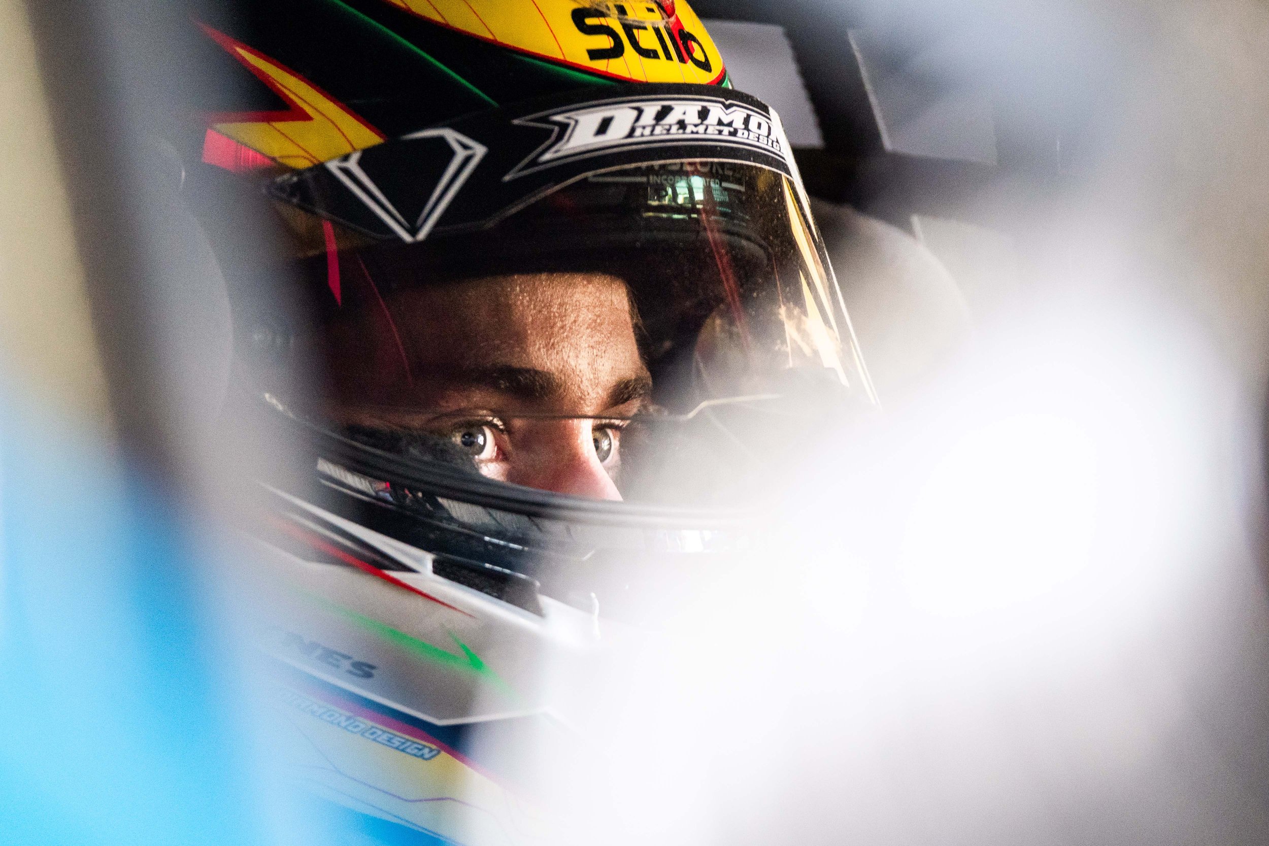Close-up of a race car driver wearing a black helmet with racing logos, looking intently.