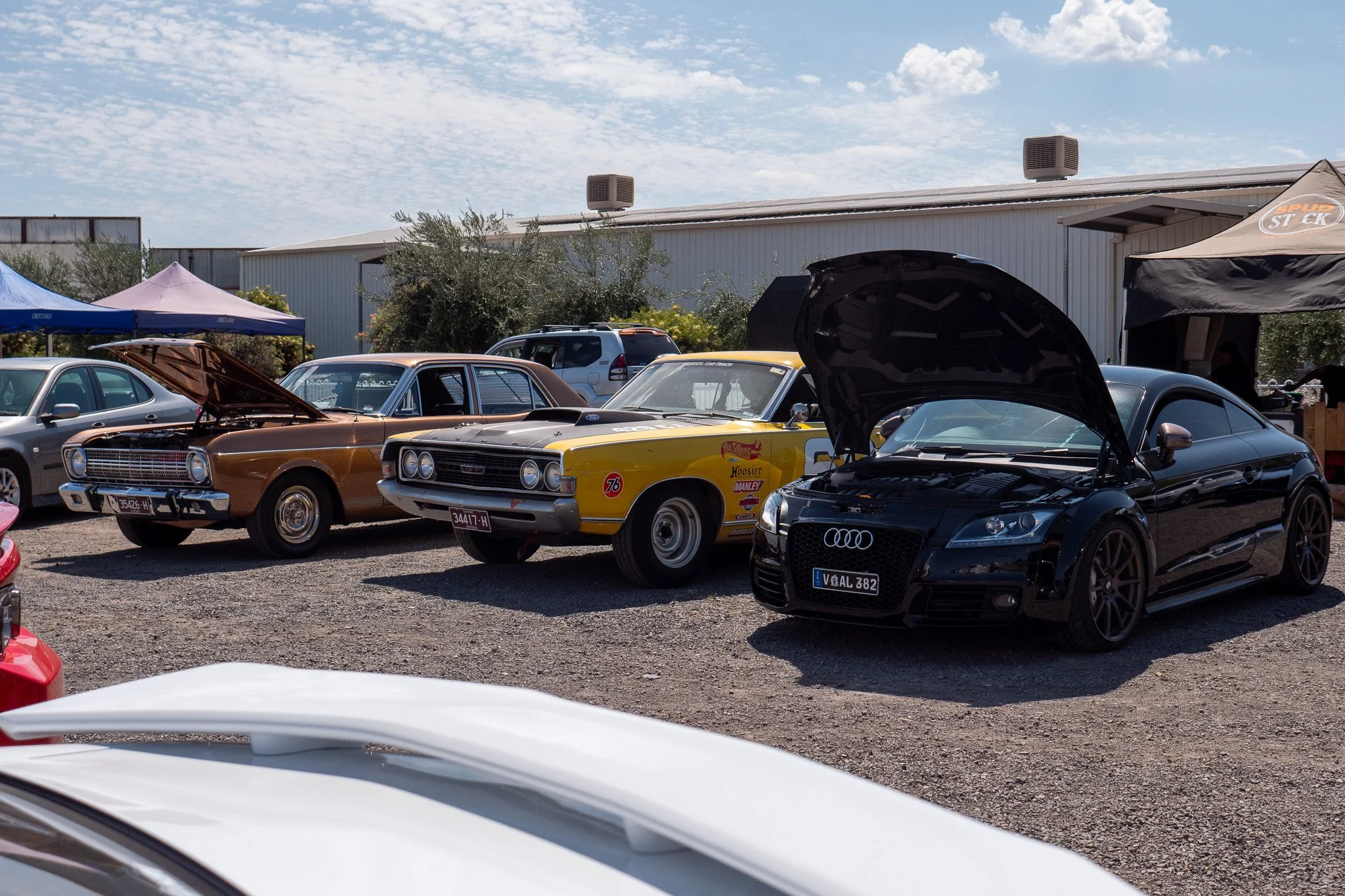 A row of classic and modern cars parked outdoors at a car show, including a black Audi with open hood, a yellow muscle car, and a brown vintage car, with tents and other vehicles in the background.