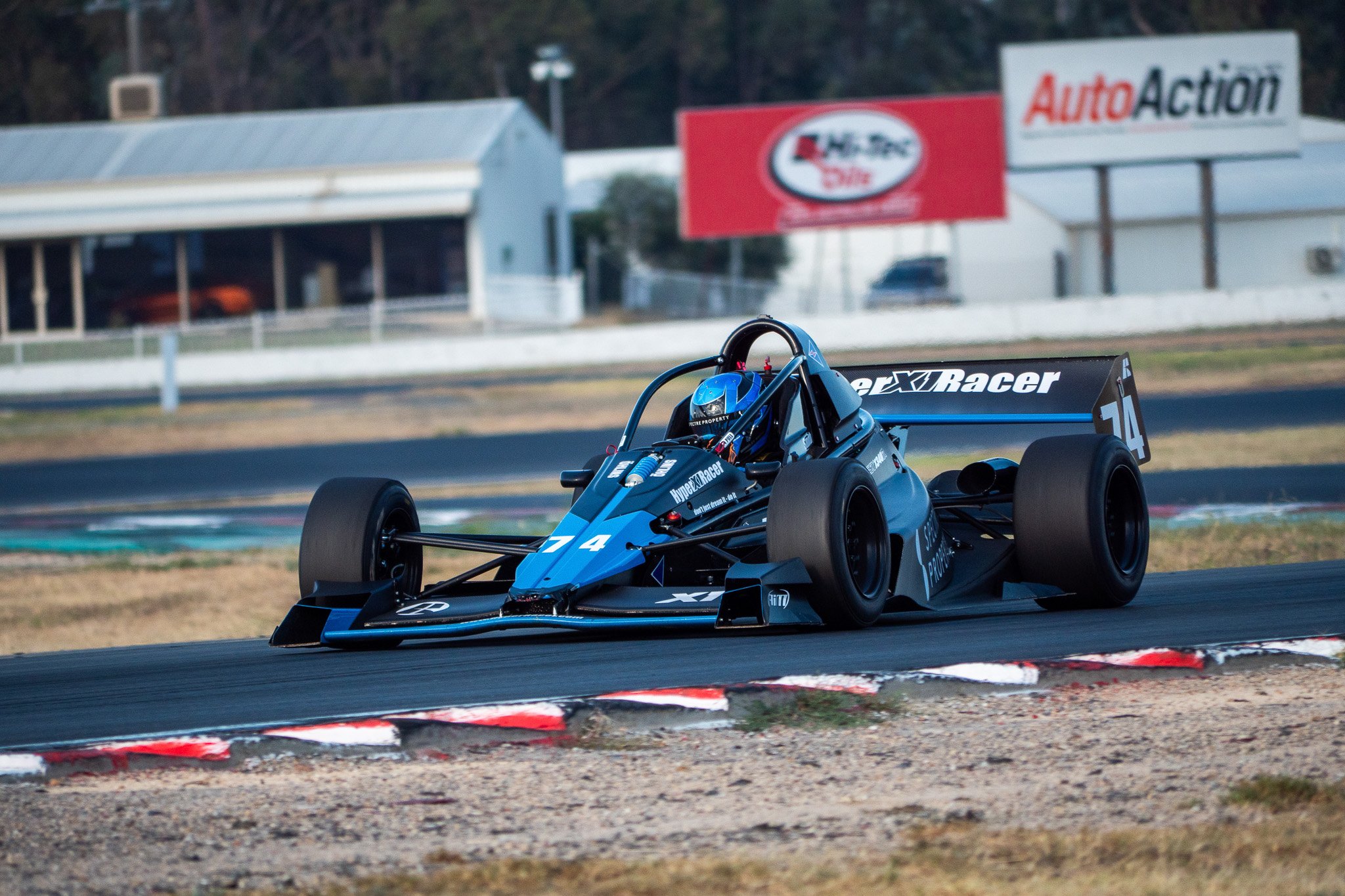 A race car with the number 14 speeding on a racetrack during daytime, with advertising signs and a building in the background.