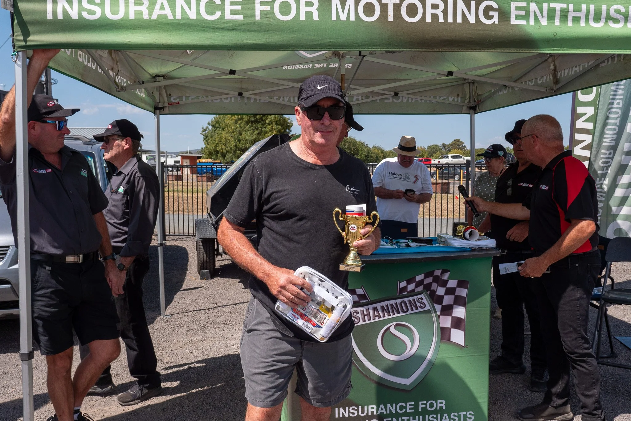 A man wearing a black hat, sunglasses, a black t-shirt, and gray shorts holding a trophy and a small toolbox at a racing event under a green canopy with a Shannon's logo and sponsor banners. Several people are in the background, some using their phon
