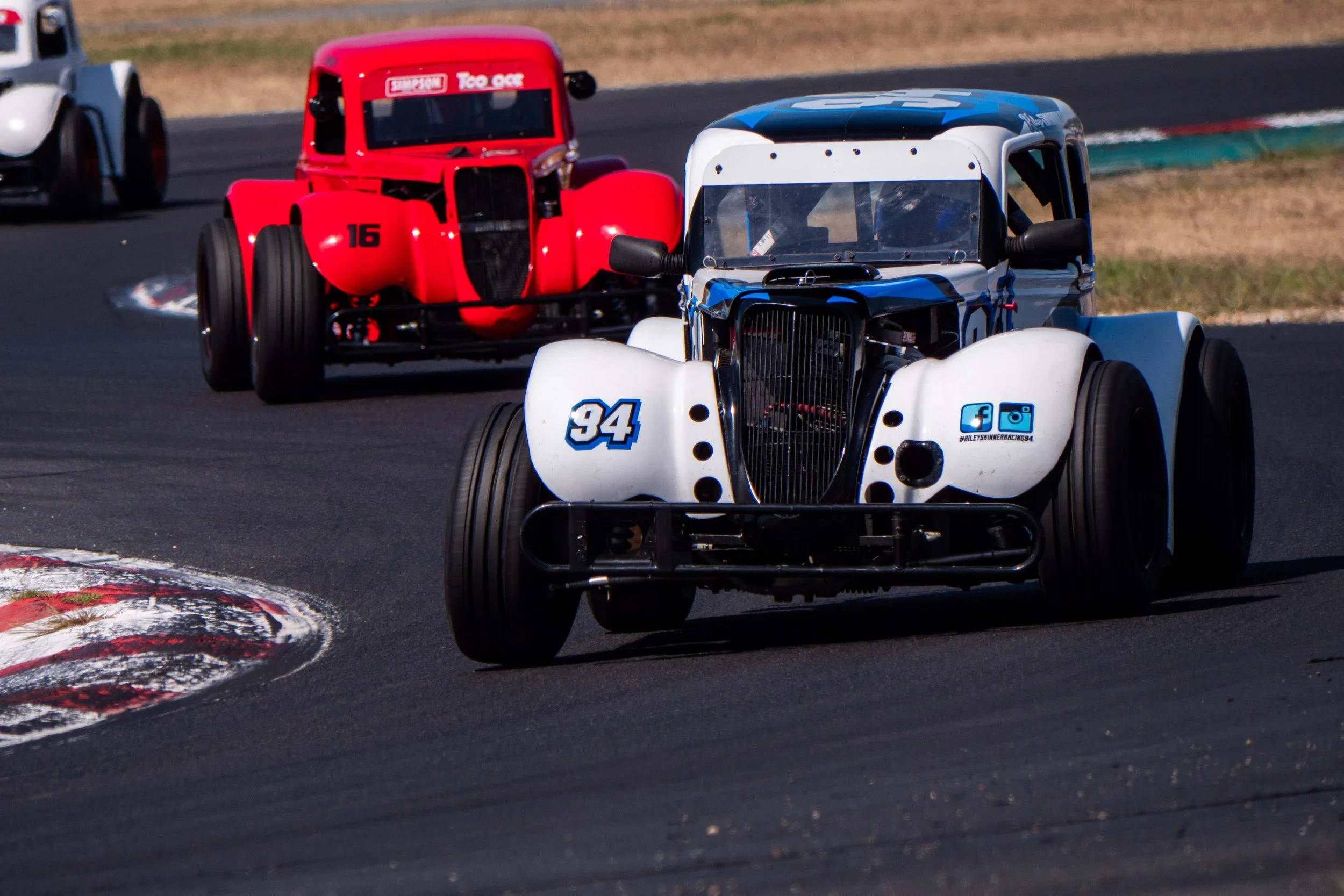 Close-up of race cars on an asphalt track, with the front white and blue car number 34 leading, followed by a red car number 16.