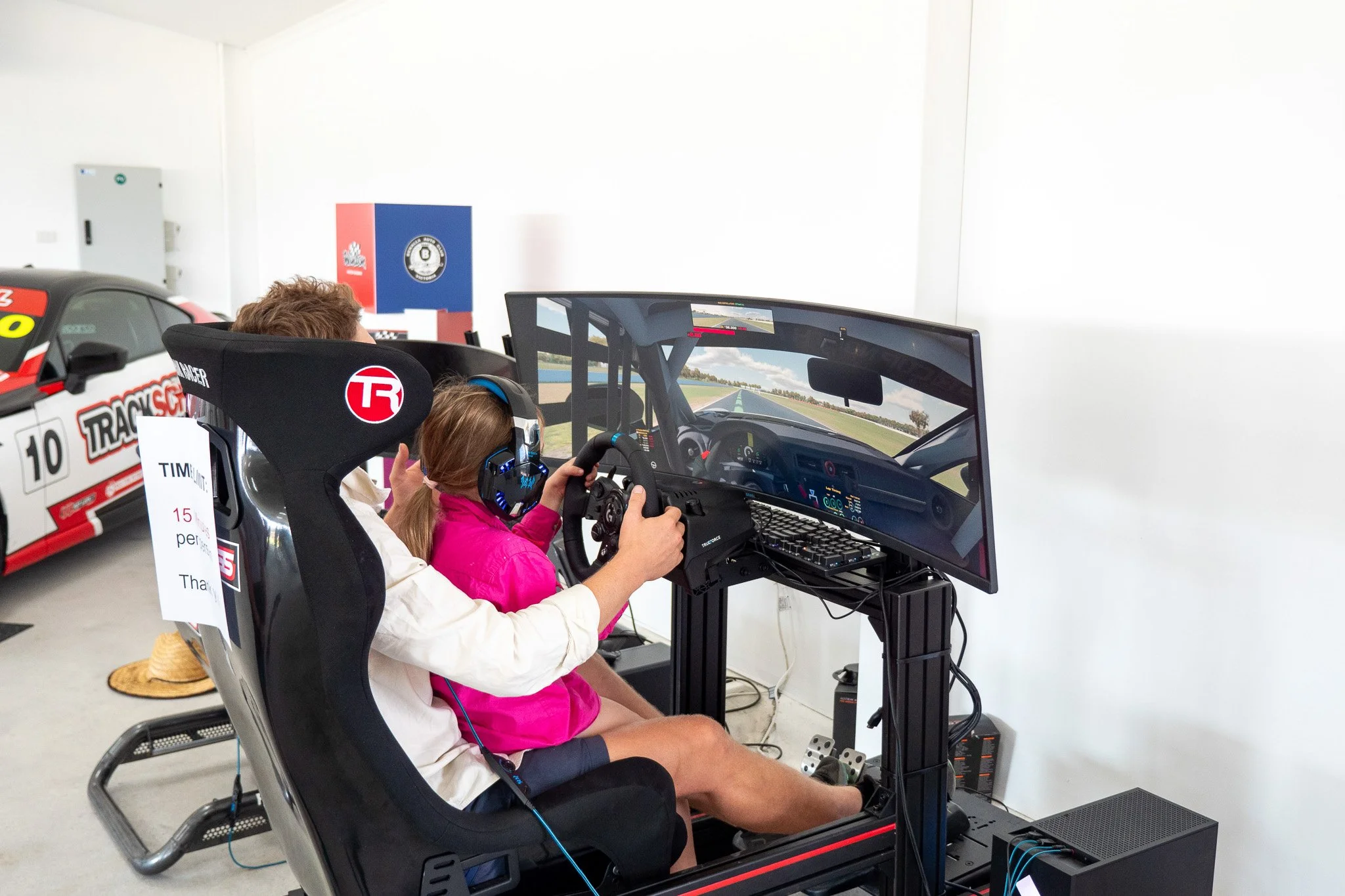 A young girl with a pink jacket and headphones sitting in a racing simulator seat riding shotgun. An adult behind her is guiding her hands on a racing wheel. There is a wide curved monitor showing a racing video game, and a racing car is visible in t