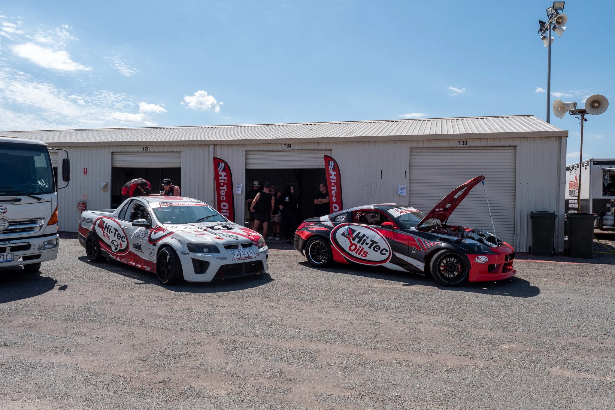 Race cars parked outside a garage with people gathering nearby. The cars are branded with 'Hi-Tec Oils'.