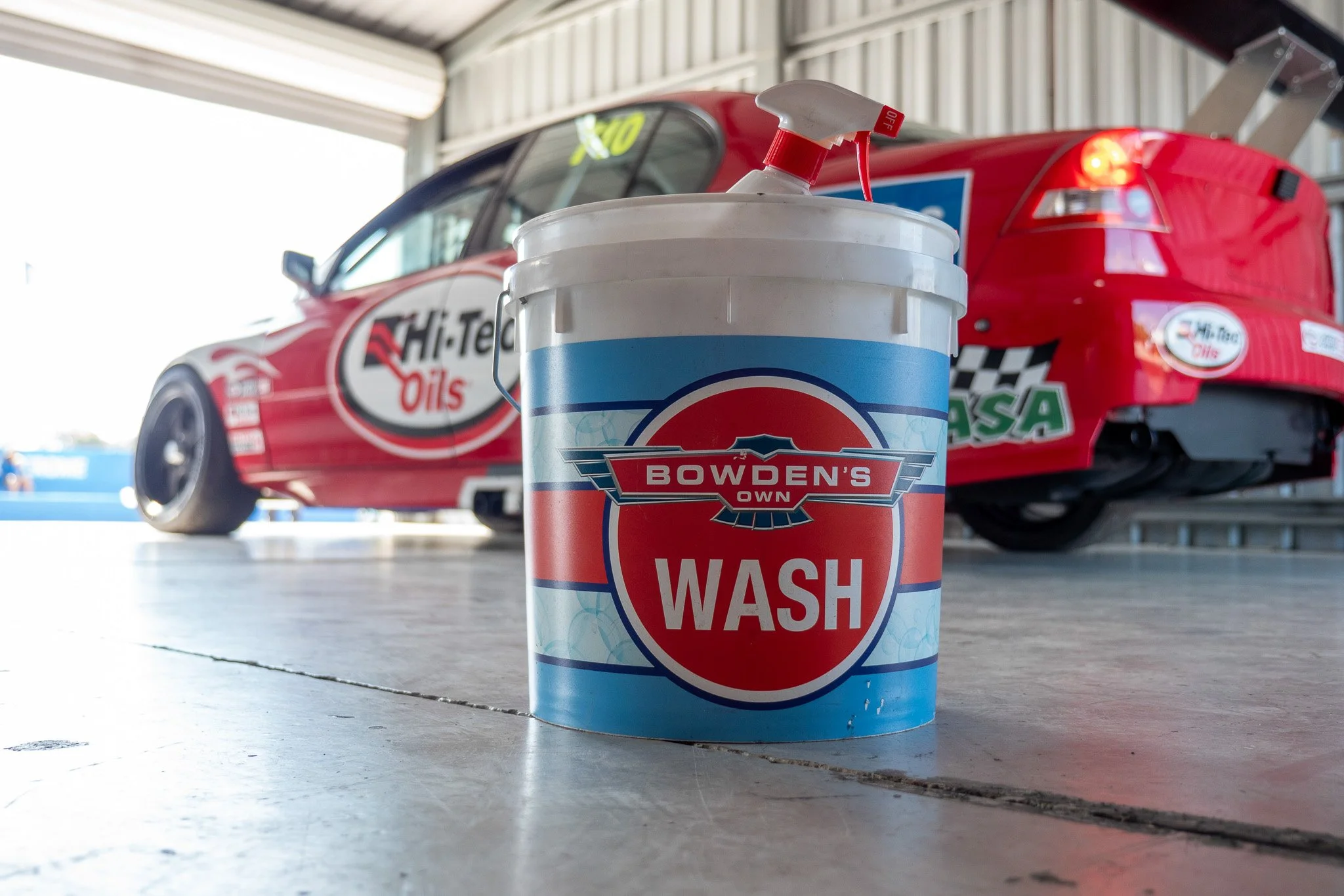 A bucket of Bowden's Own Wash in front of a red race car with Hi-Tech Oils branding.