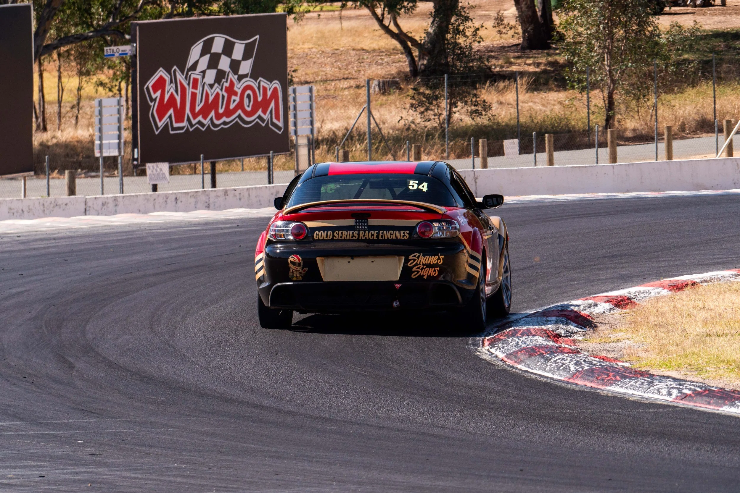 A race car on a track, captured from the rear, taking a turn with a Wilton billboard in the background.