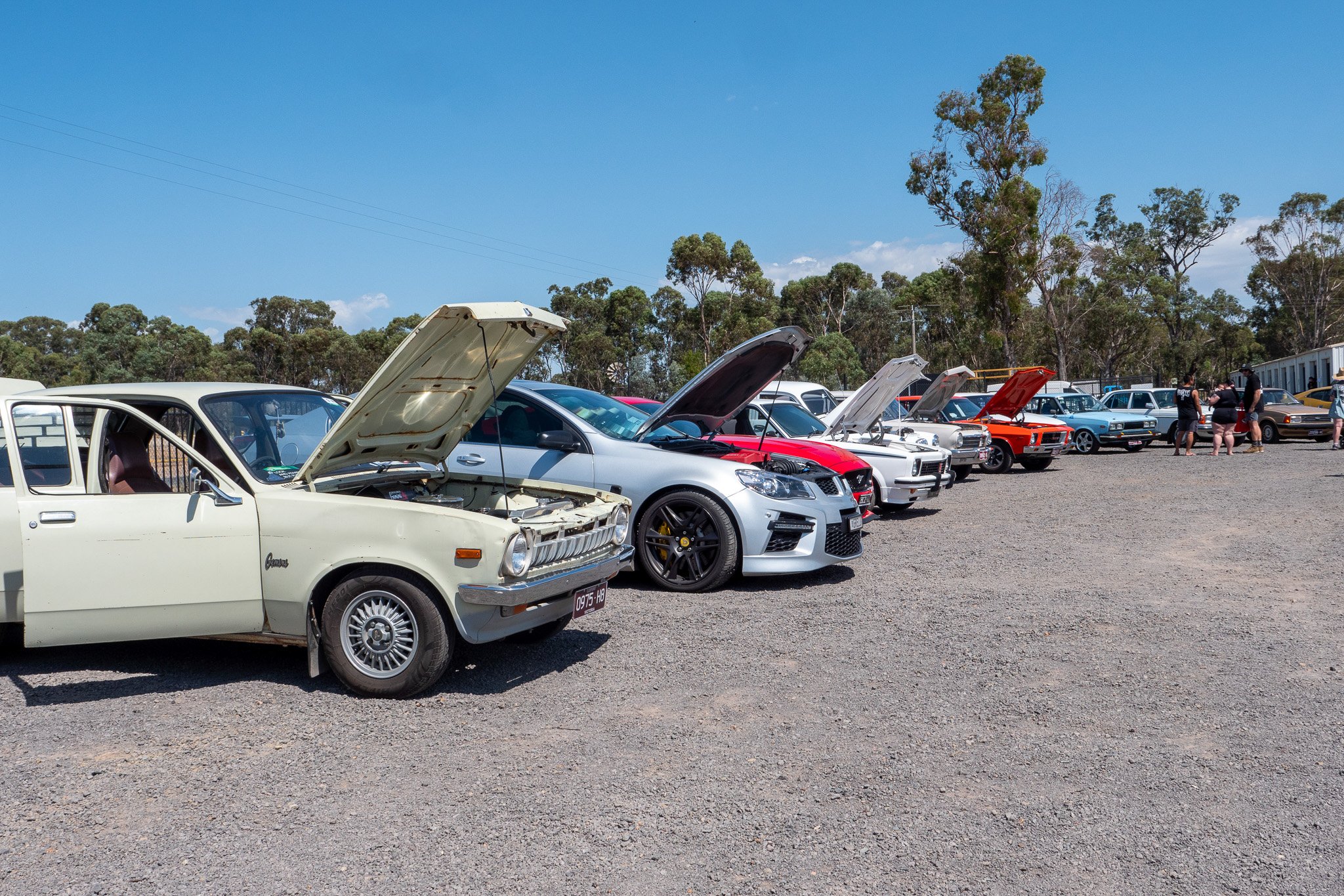 A lineup of vintage and modern cars at a car show, with some cars' hoods open for viewing, on a gravel lot under a clear blue sky, with trees and a few people in the background.