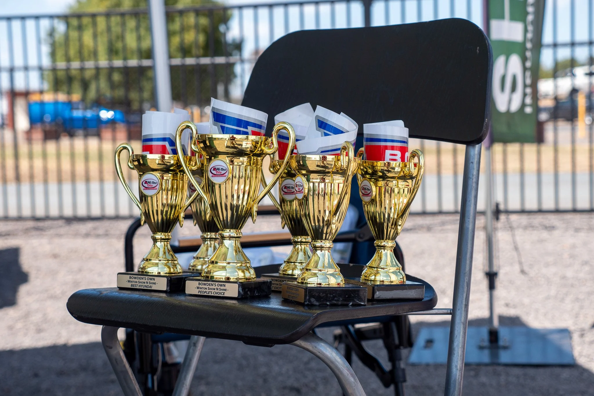 Four gold trophies placed on a black chair outdoors, with small banners and trees in the background.