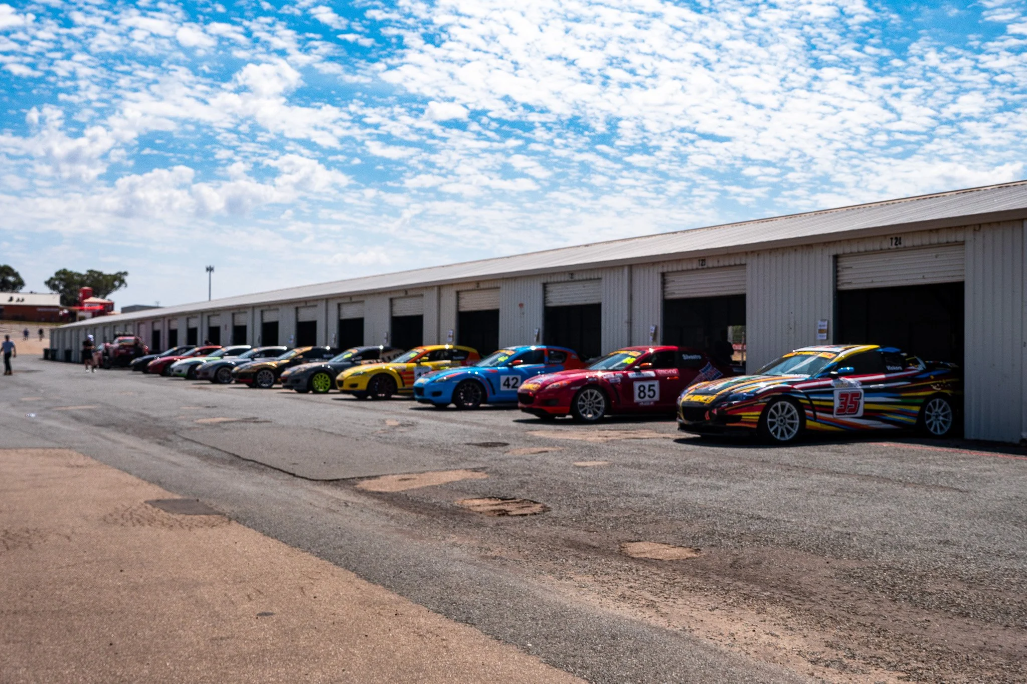 Multiple race cars parked in front of a garage at the race track, with a partly cloudy sky overhead.