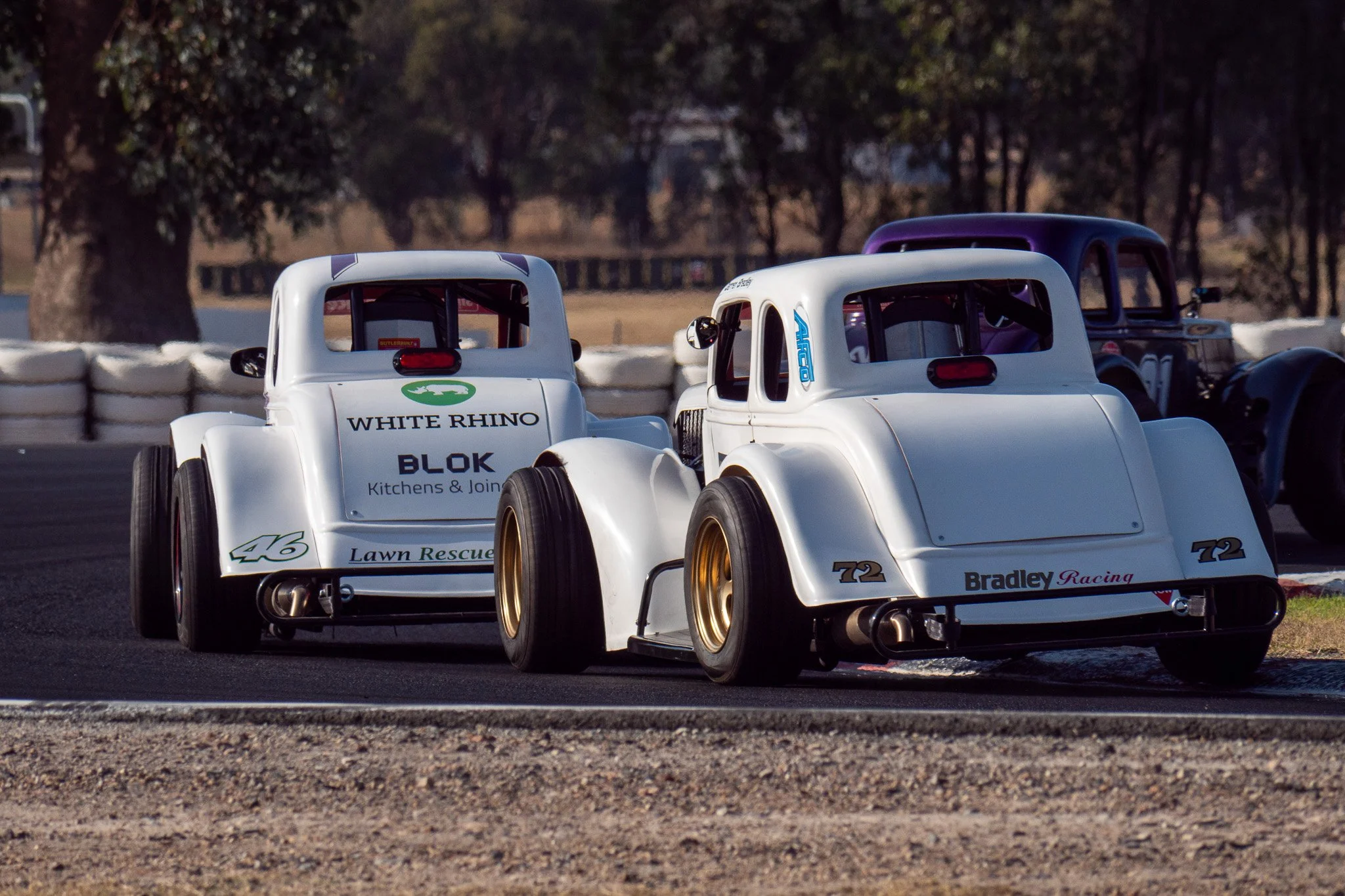 Two vintage race cars, white with gold wheels, racing on a track with a third car partially visible behind them.