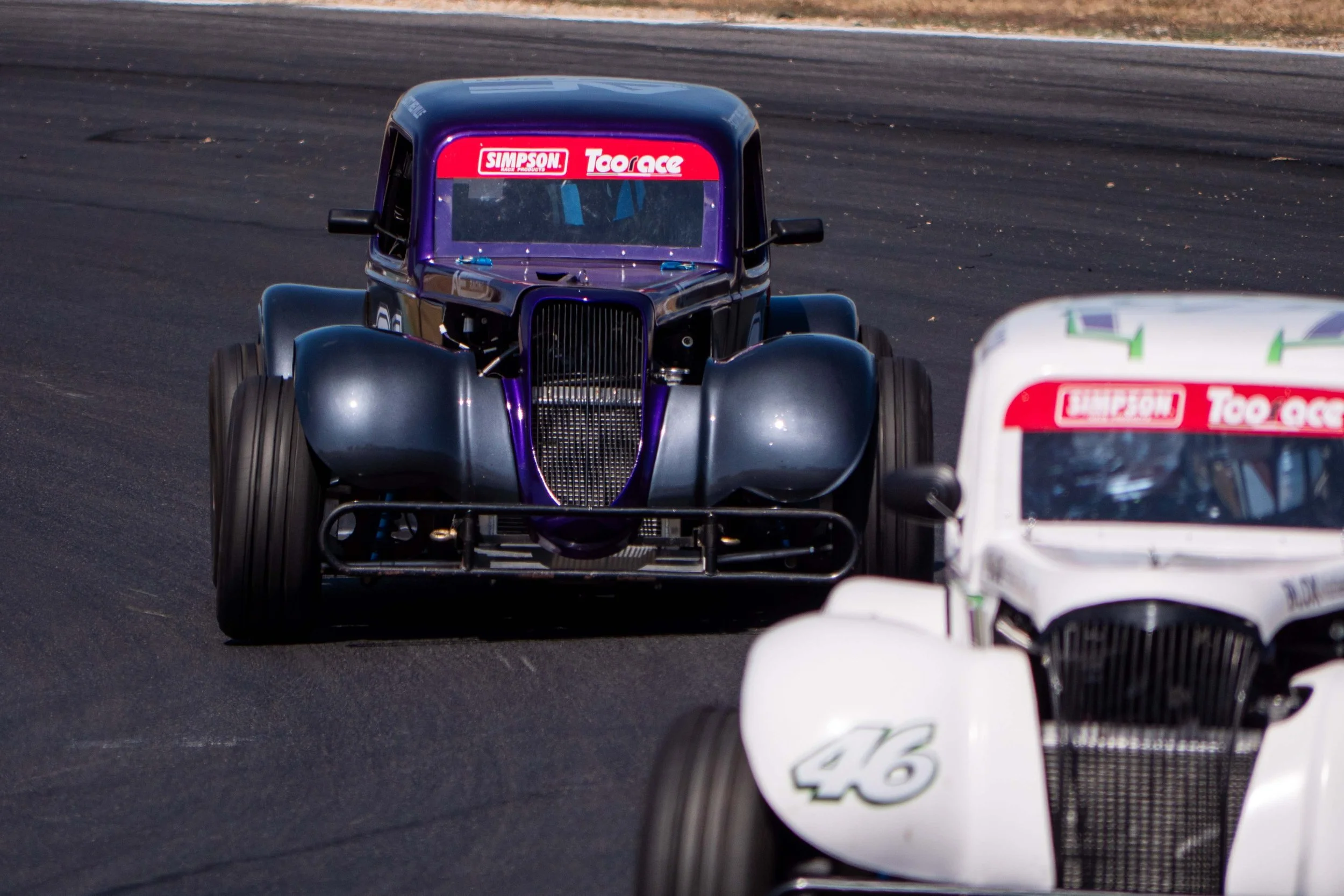 Two vintage race cars on a racetrack, one black with purple accents and the other white, both featuring sponsor decals and racing numbers.