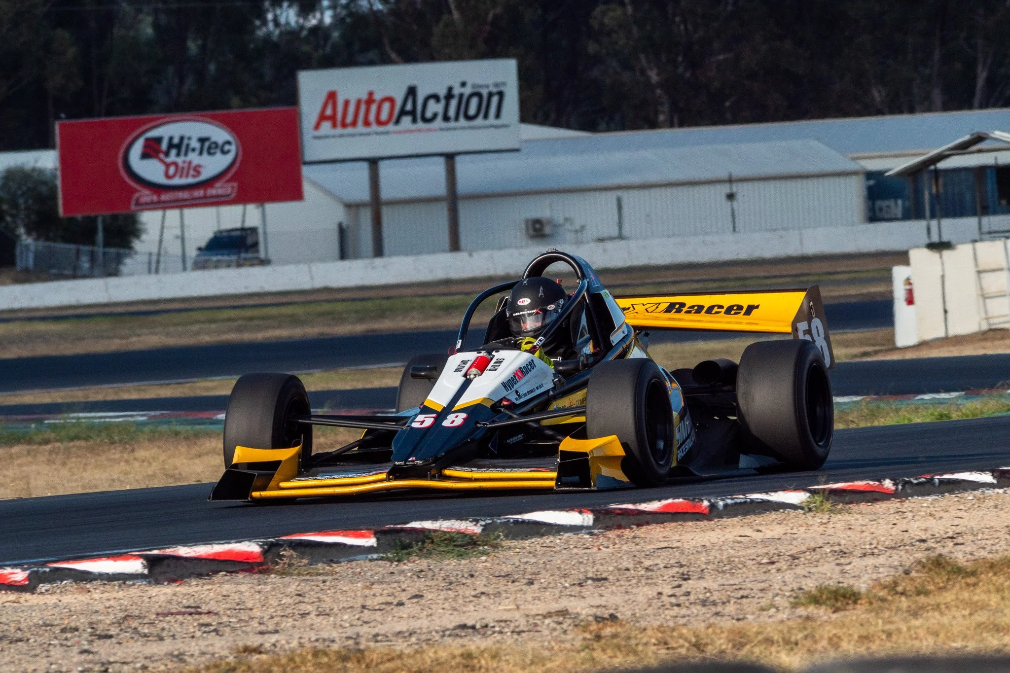 A yellow and black open-wheel race car on a race track, with a driver wearing a black helmet, around a corner during a race.