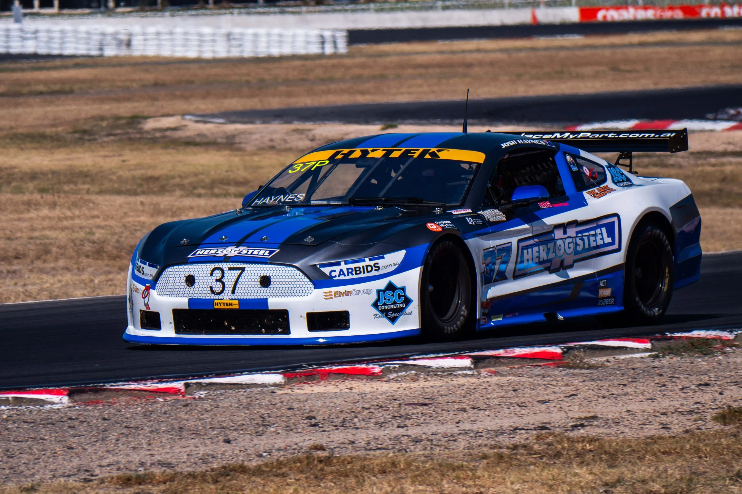 A blue and white race car with the number 37 on the front, driving on a racetrack with a black and red curb.