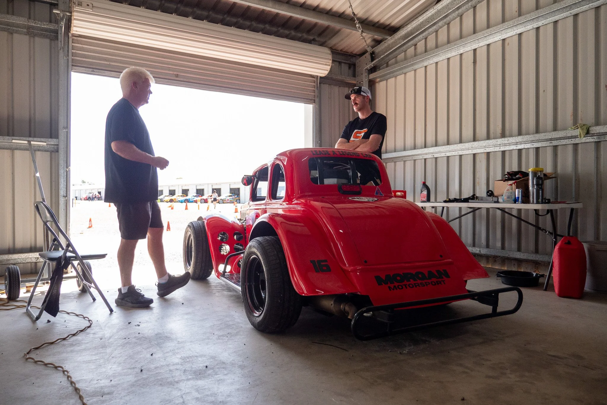 Two men standing inside a garage with a bright open door, next to a red race car. One man has platinum blonde hair and is wearing a black T-shirt and shorts. The other man is wearing a cap and a black T-shirt with an orange and white logo, smiling. T
