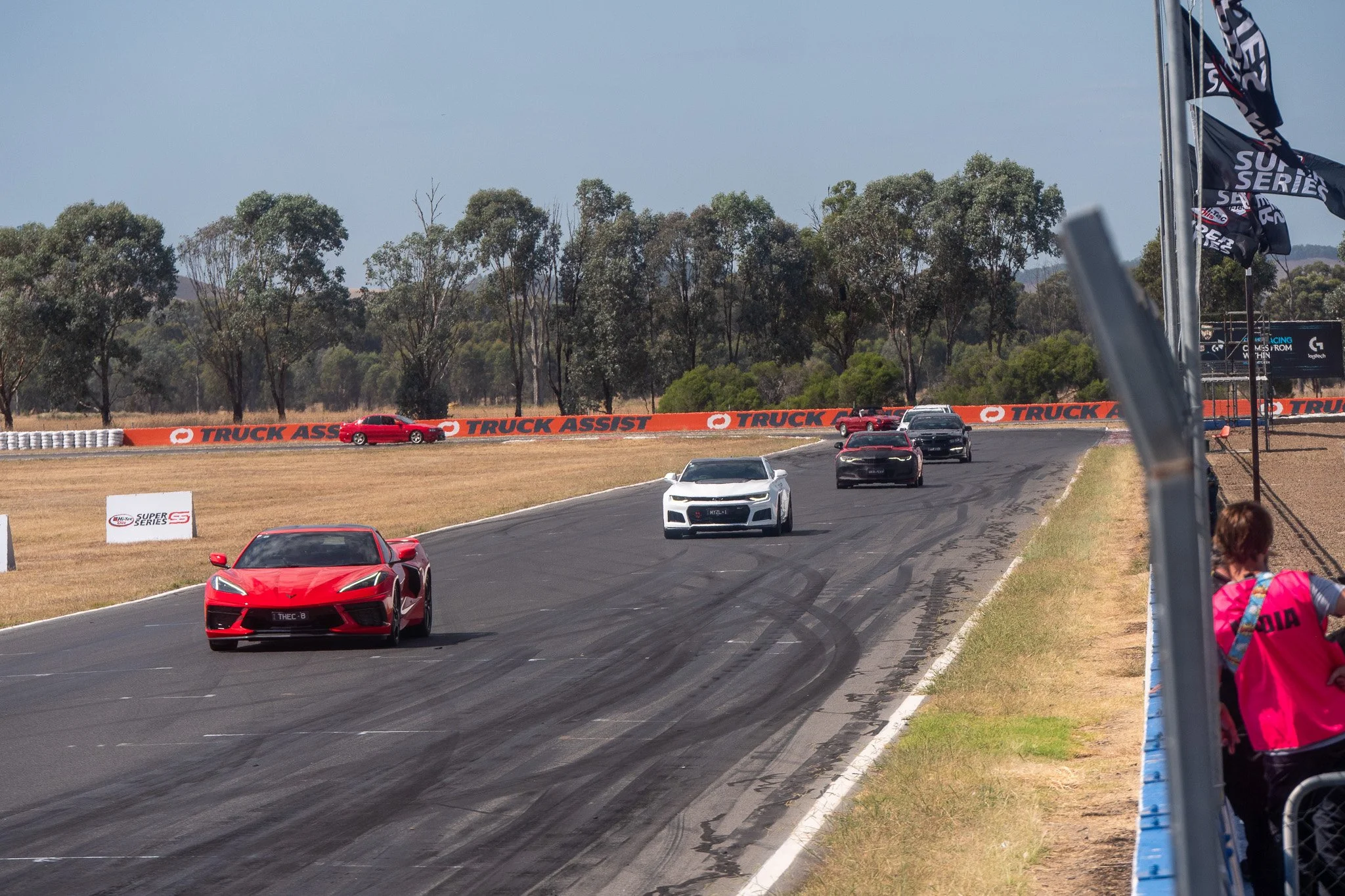 Multiple race cars on a dirt race track during a motorsport event, with trees and banners in the background.