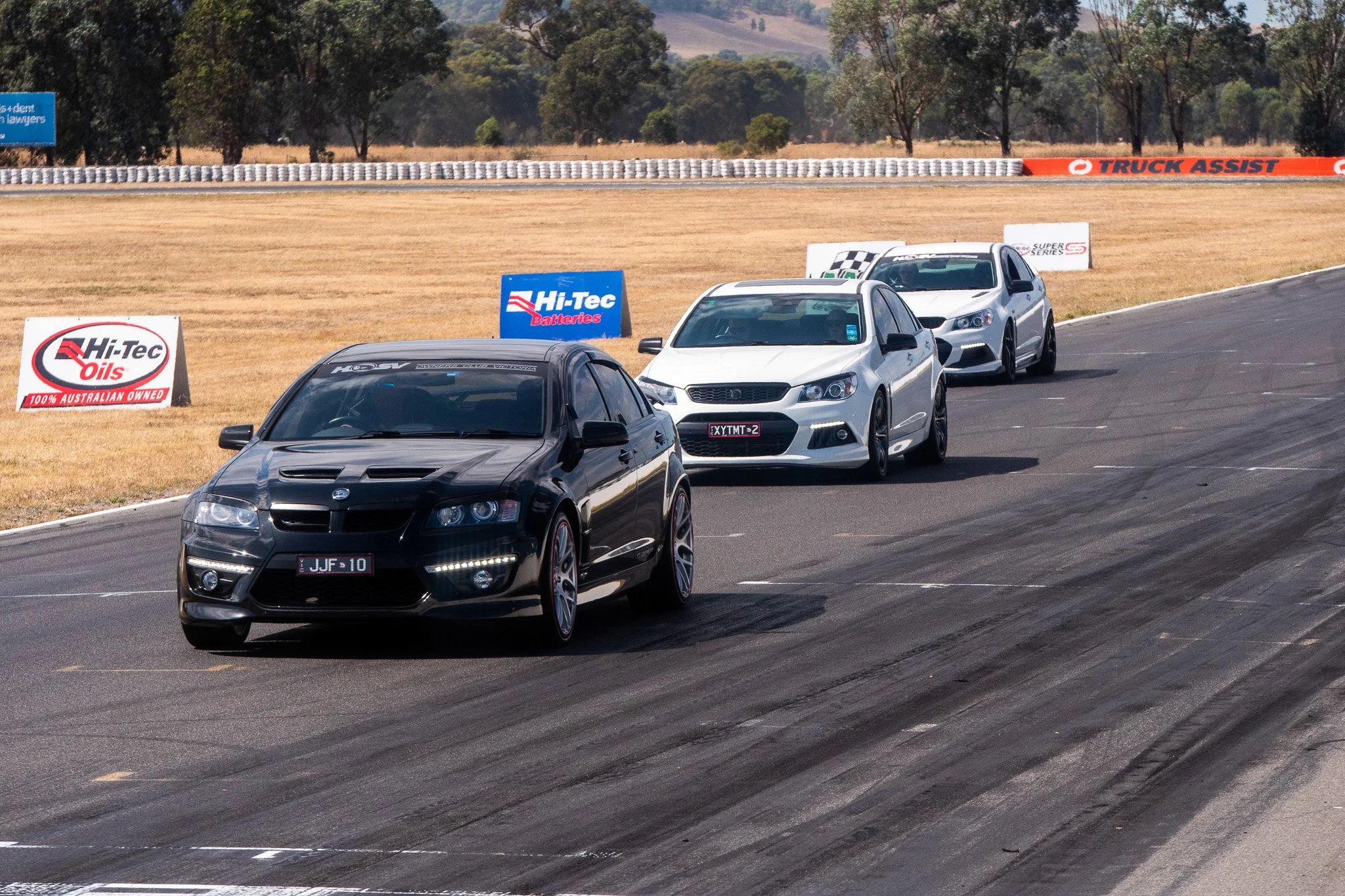 Three cars racing on a track with grass in the background and advertising banners along the side of the track.
