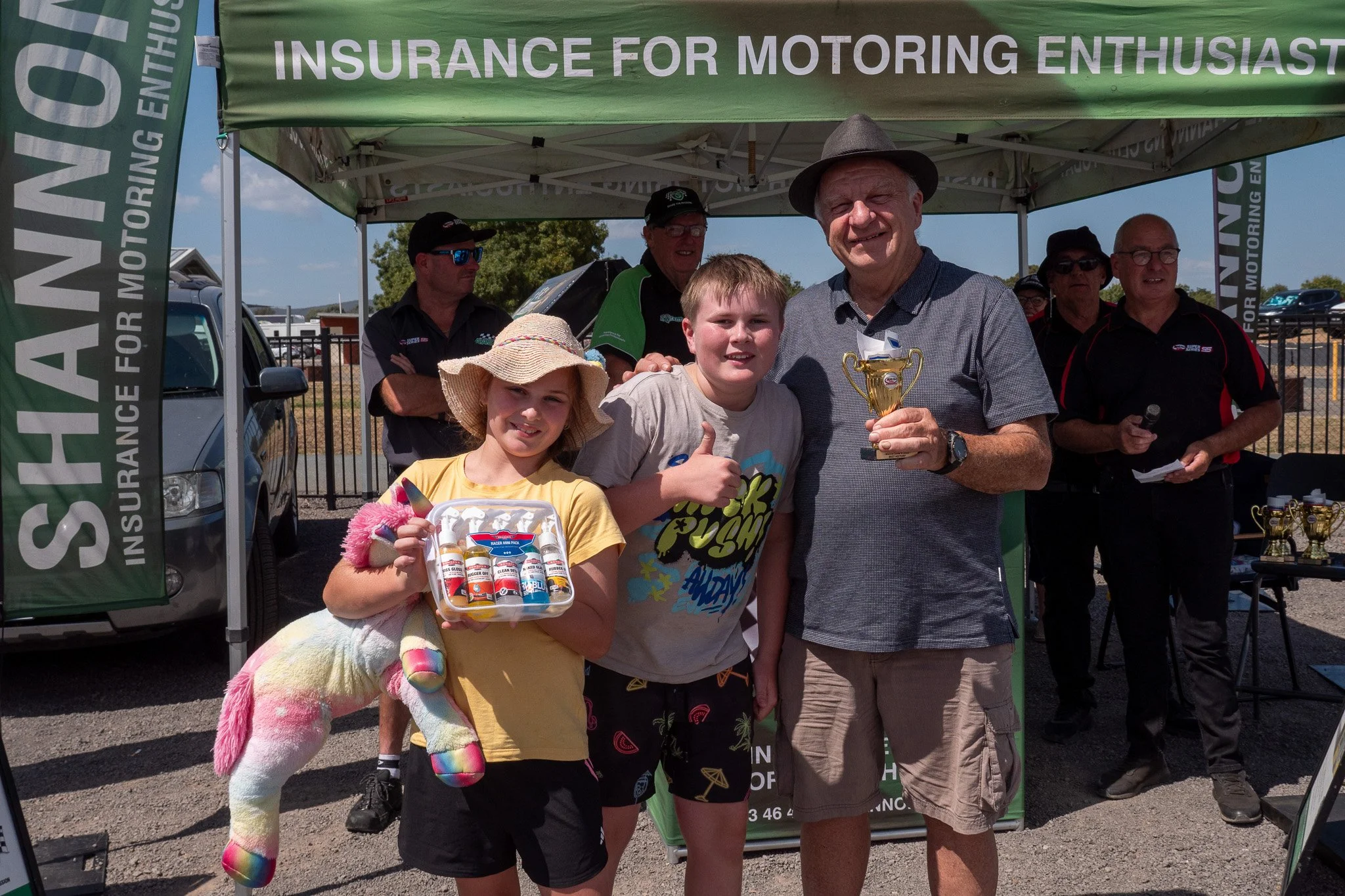 People standing under a green tent at an event, one man holding a trophy, children, and adults smiling and posing for the photo.
