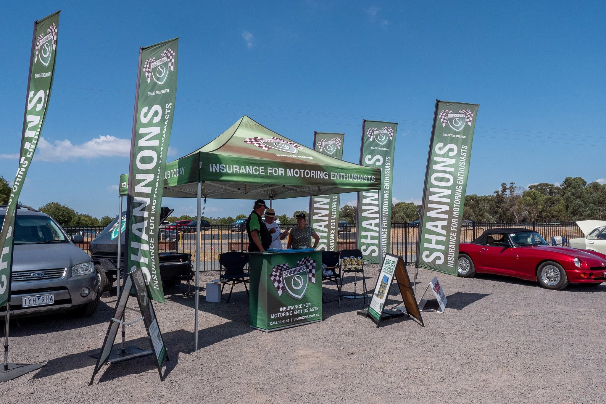 Outdoor event booth with green tent and flags advertising insurance for motoring enthusiasts, surrounded by parked cars on a sunny day.