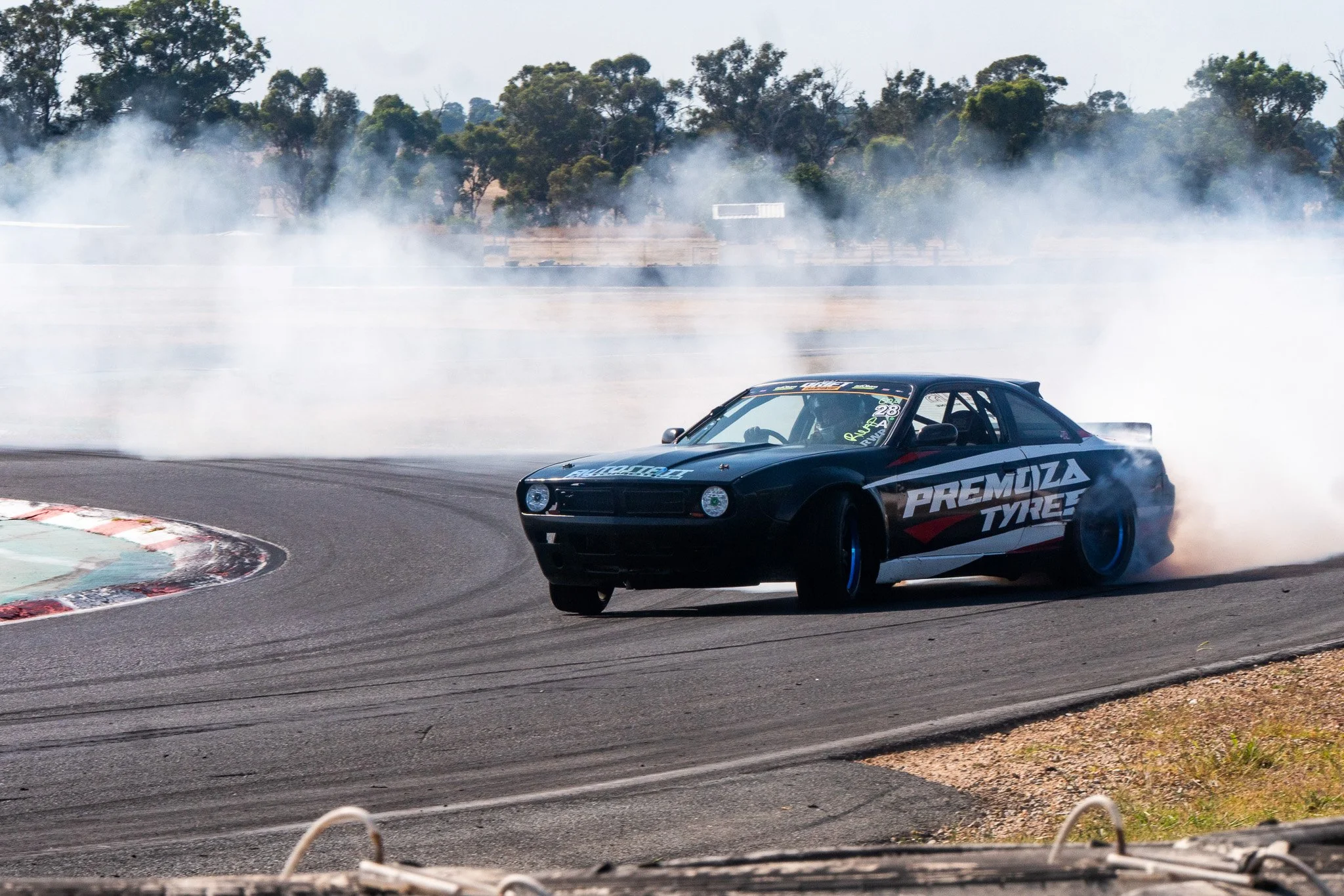 A black race car with sponsorship logos, including Premiza Tyre, appears to be drifting on a race track, creating tire smoke as it navigates a turn, with trees and a clear sky in the background.