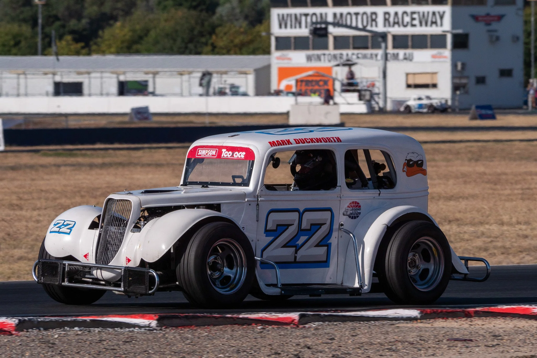 A vintage race car with the number 22 is on a racetrack at Winton Motor Raceway, with a driver in racing gear inside. The car is white with decals, and the background shows race facilities and signage.