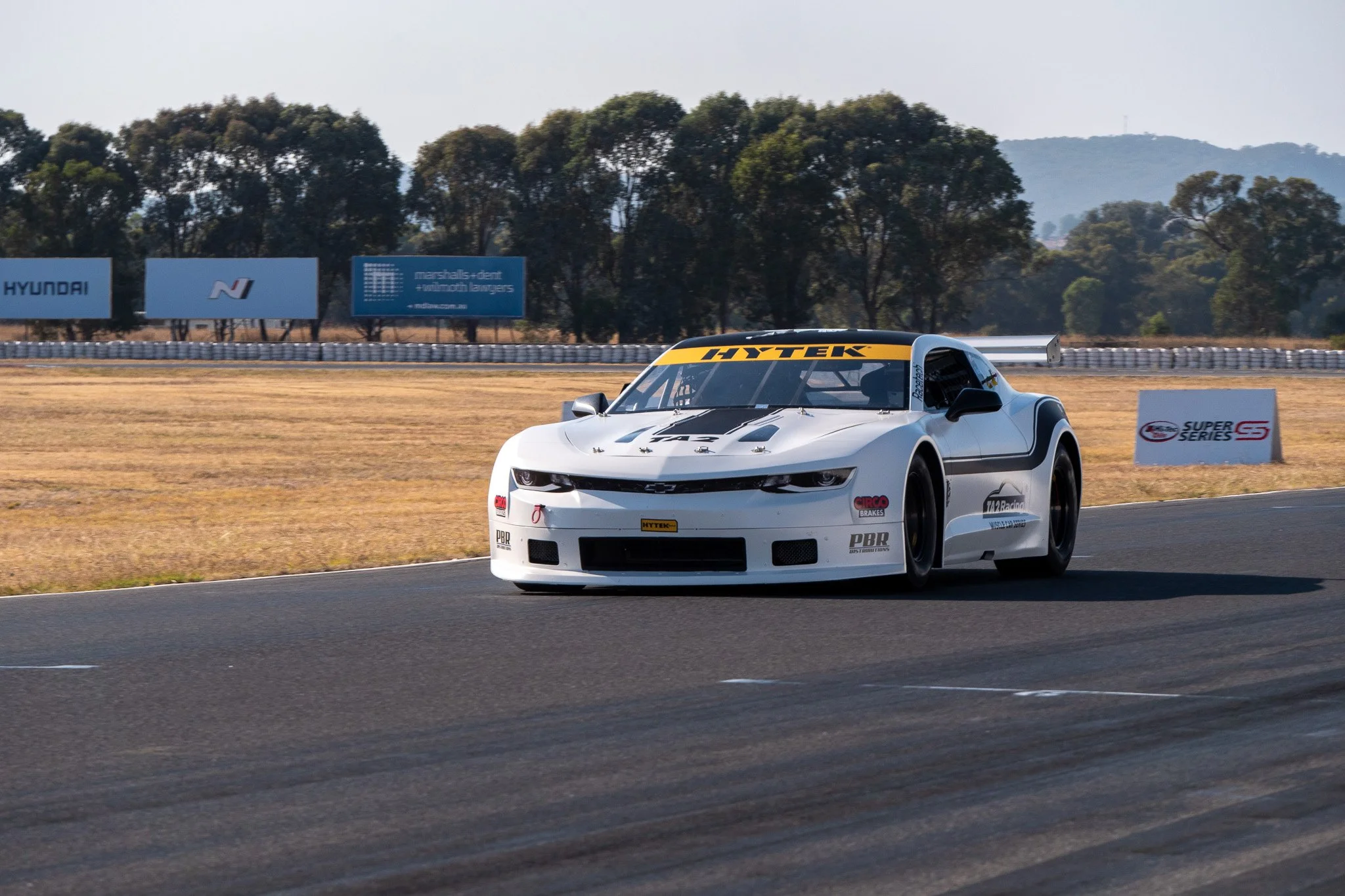 A white race car on a racetrack with trees and billboards in the background during daytime.