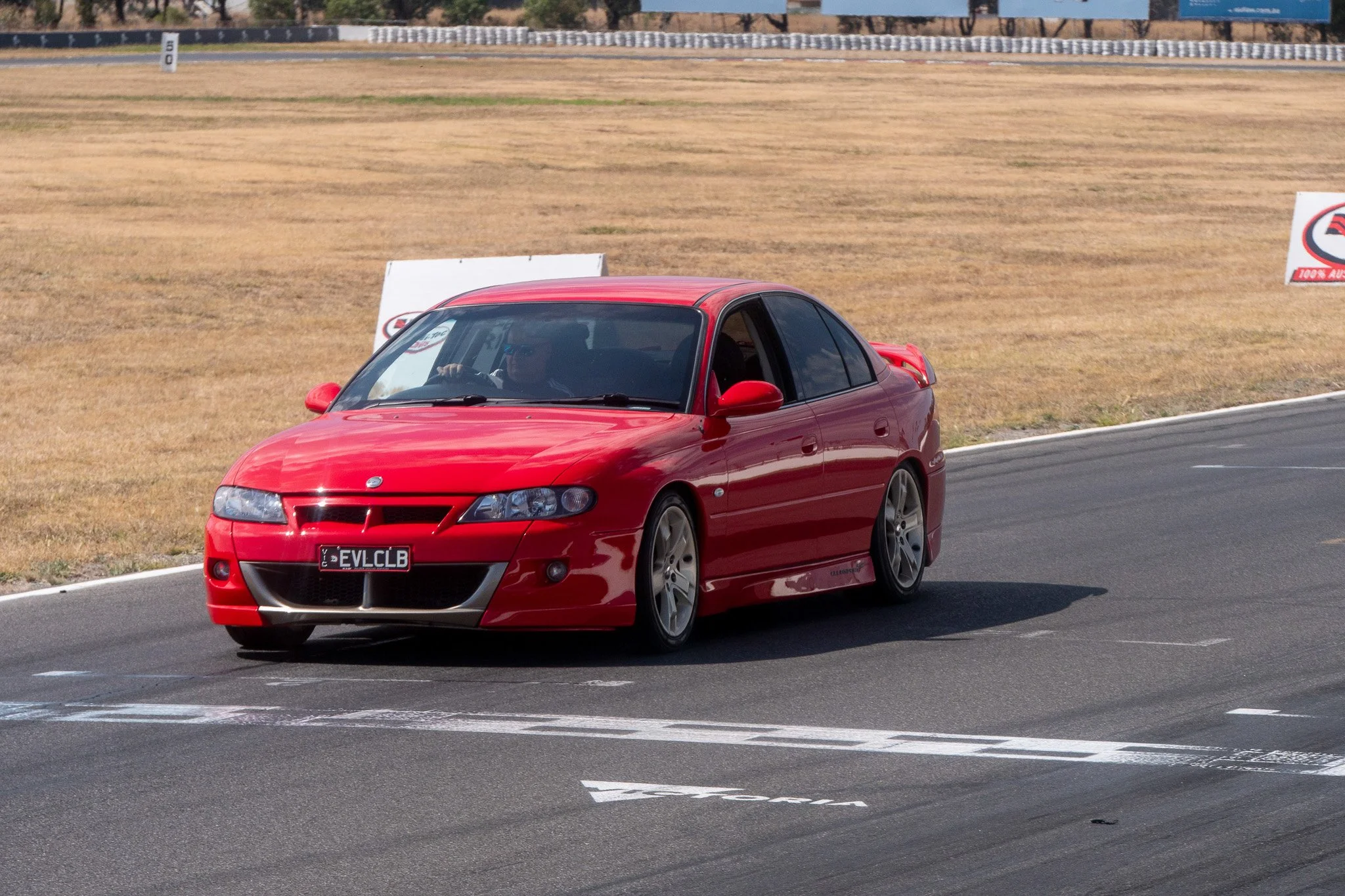 A red sedan car on a race track during daytime, with a driver and a passenger inside, and track markings visible on the pavement.