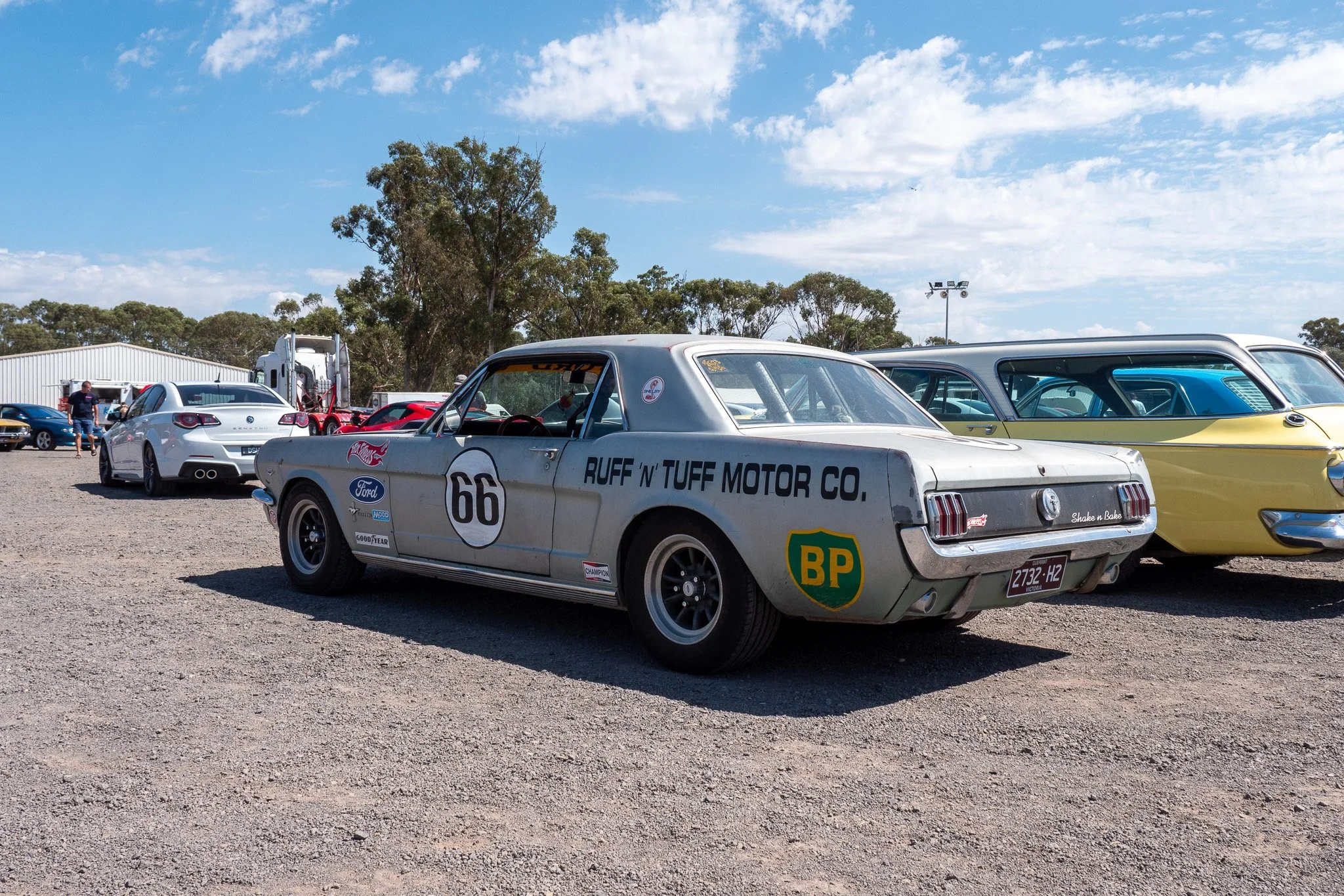 A vintage Ford race car with the number 66 and various sponsor logos parked on a gravel lot alongside other classic cars, with a clear blue sky and trees in the background.