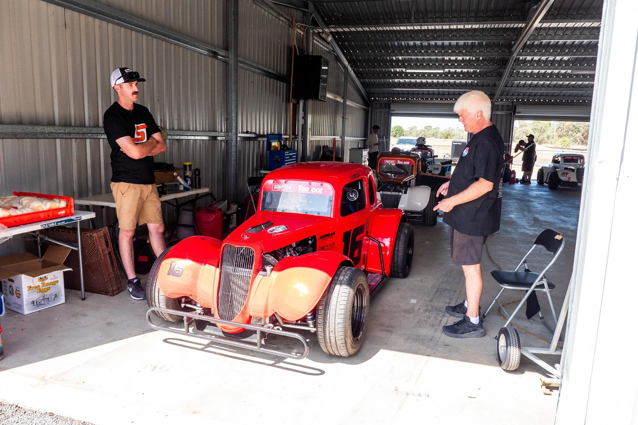 Two men in a garage with vintage race cars. One is wearing a black shirt and tan shorts, the other in a black shirt and shorts. The red race car with the number 16 is in the center, with other cars in the background.
