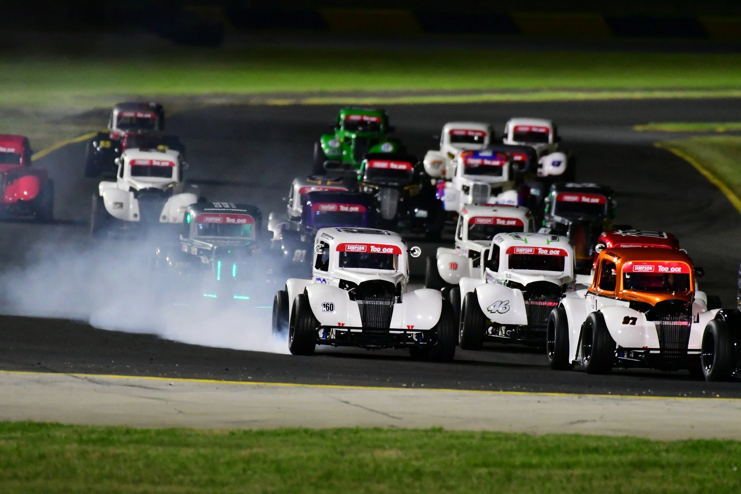 Nighttime car race featuring vintage-style race cars on a track, with some cars emitting tire smoke.