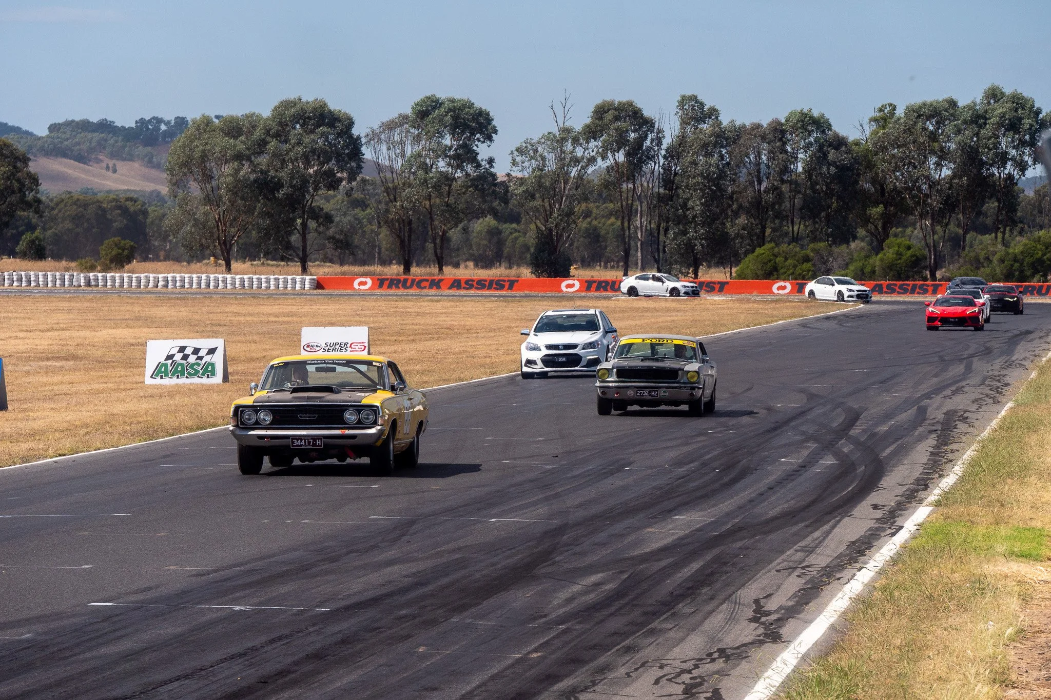 Several vintage and modern cars racing on a track with tire marks, surrounded by grass and trees, with a backdrop of hills and a clear sky.