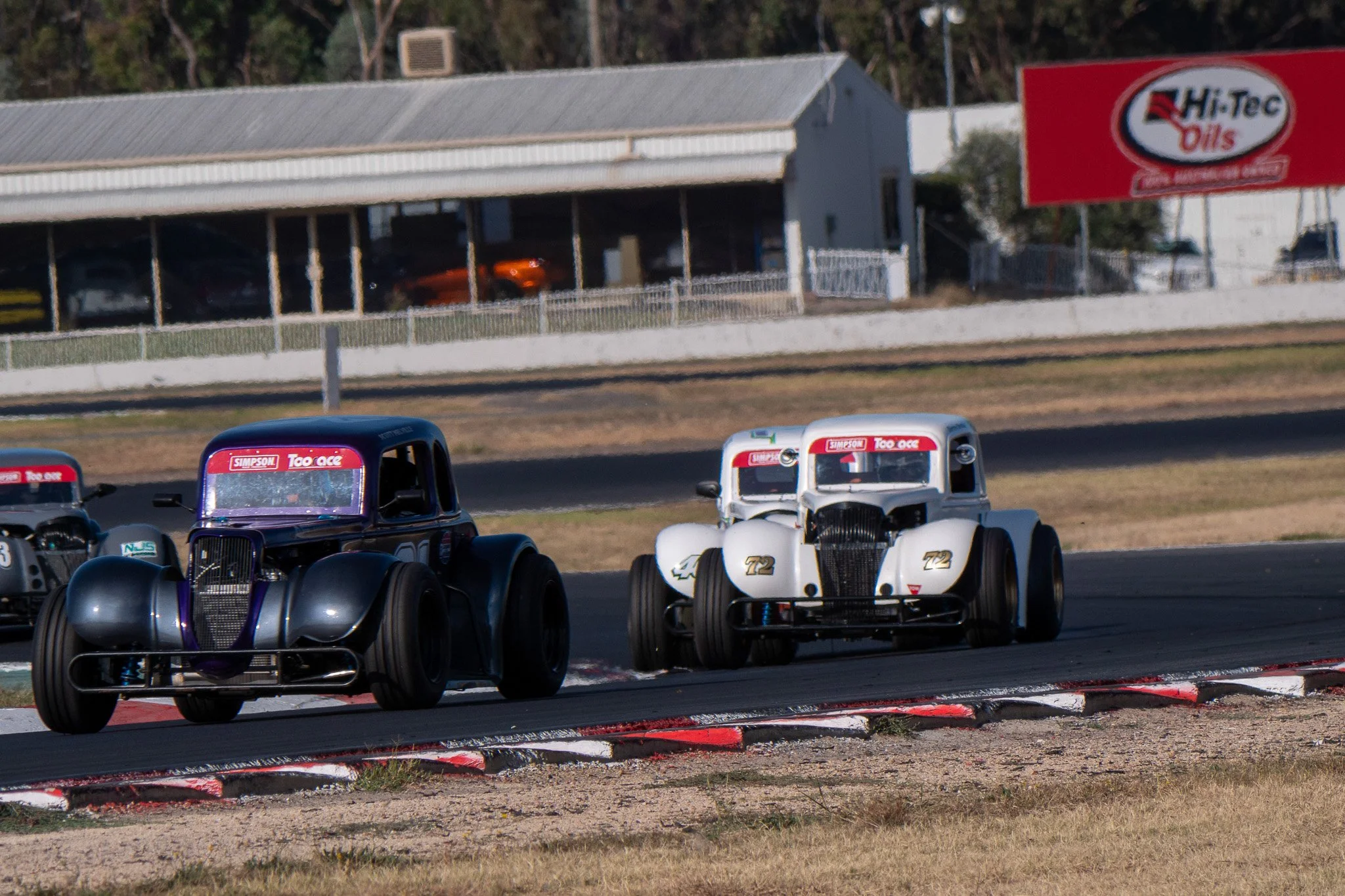 Vintage race cars on a track during a race, with a building and sign in the background.