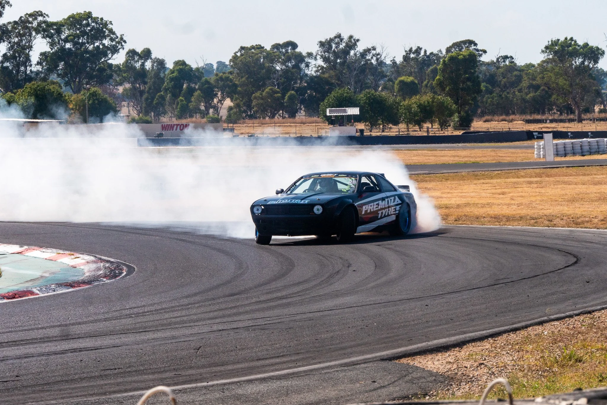 A race car drifting on a race track with smoke coming from the rear tires, surrounded by a rural landscape with trees and fencing in the background.