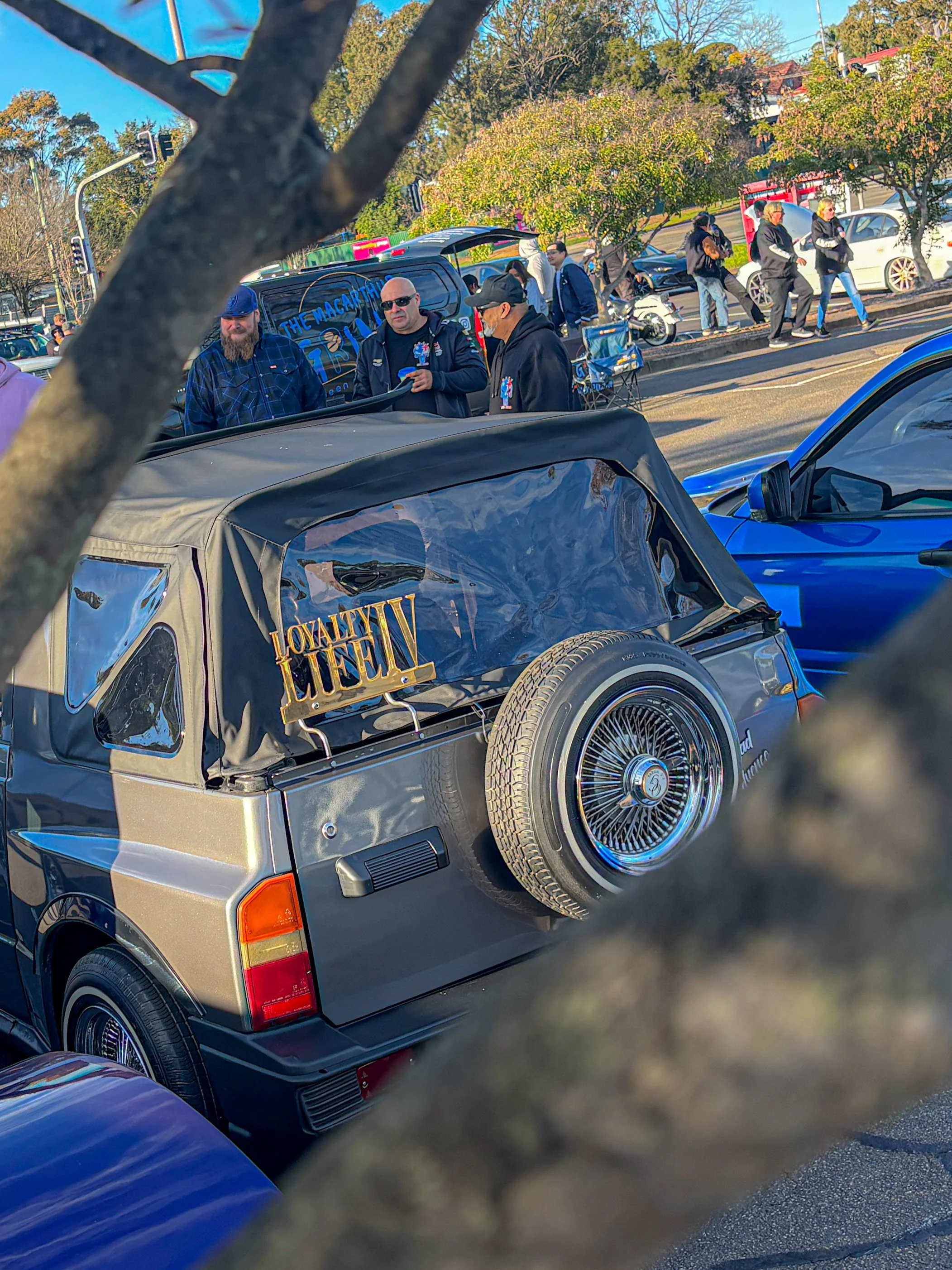 A vintage silver convertible car with a spare tire mounted on the back, partially obscured by a tree branch in the foreground. People are gathered around in the background, some conversing, near a street with a blue car parked adjacent.