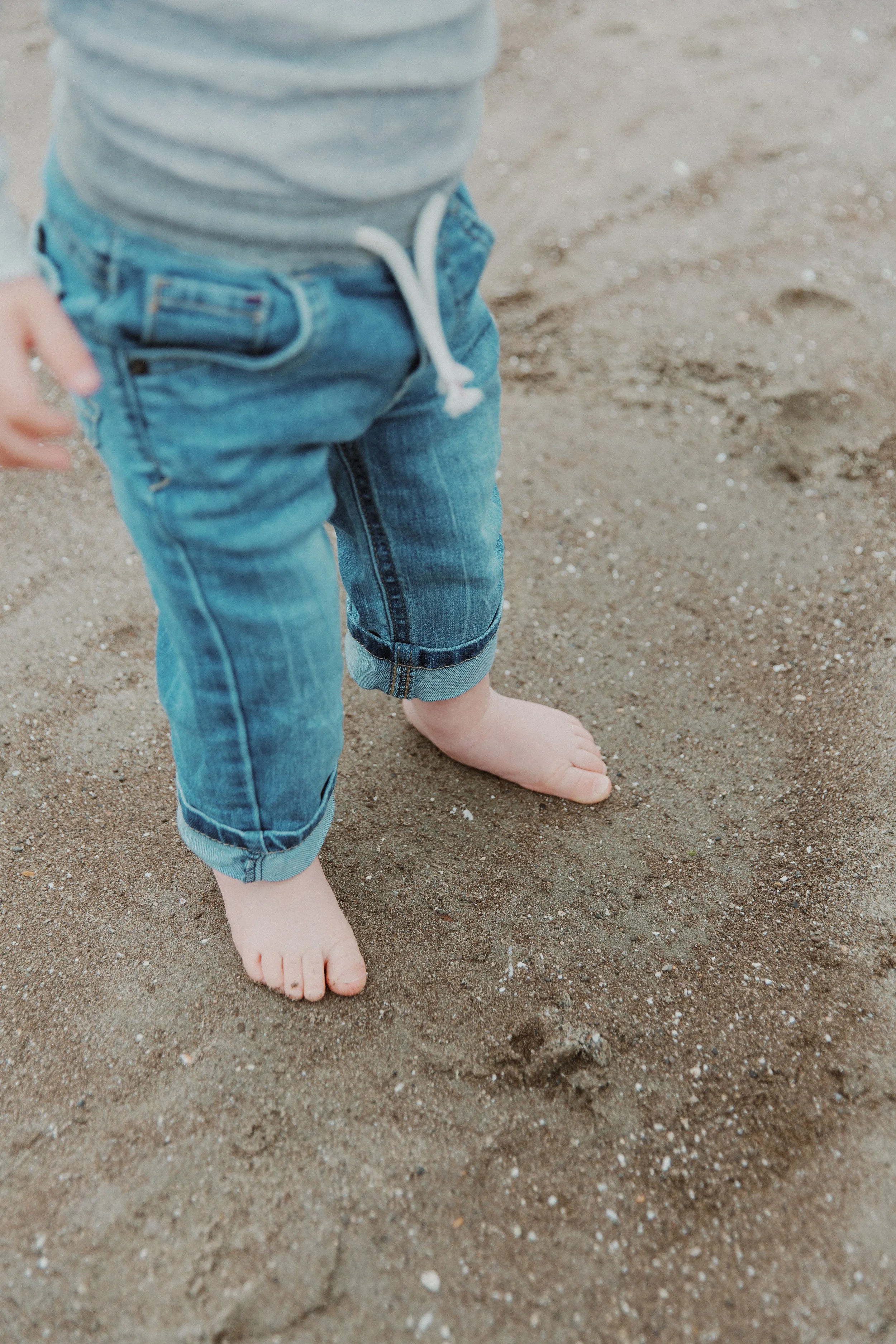 Close-up of a toddler's bare feet in the sand wearing rolled-up jeans