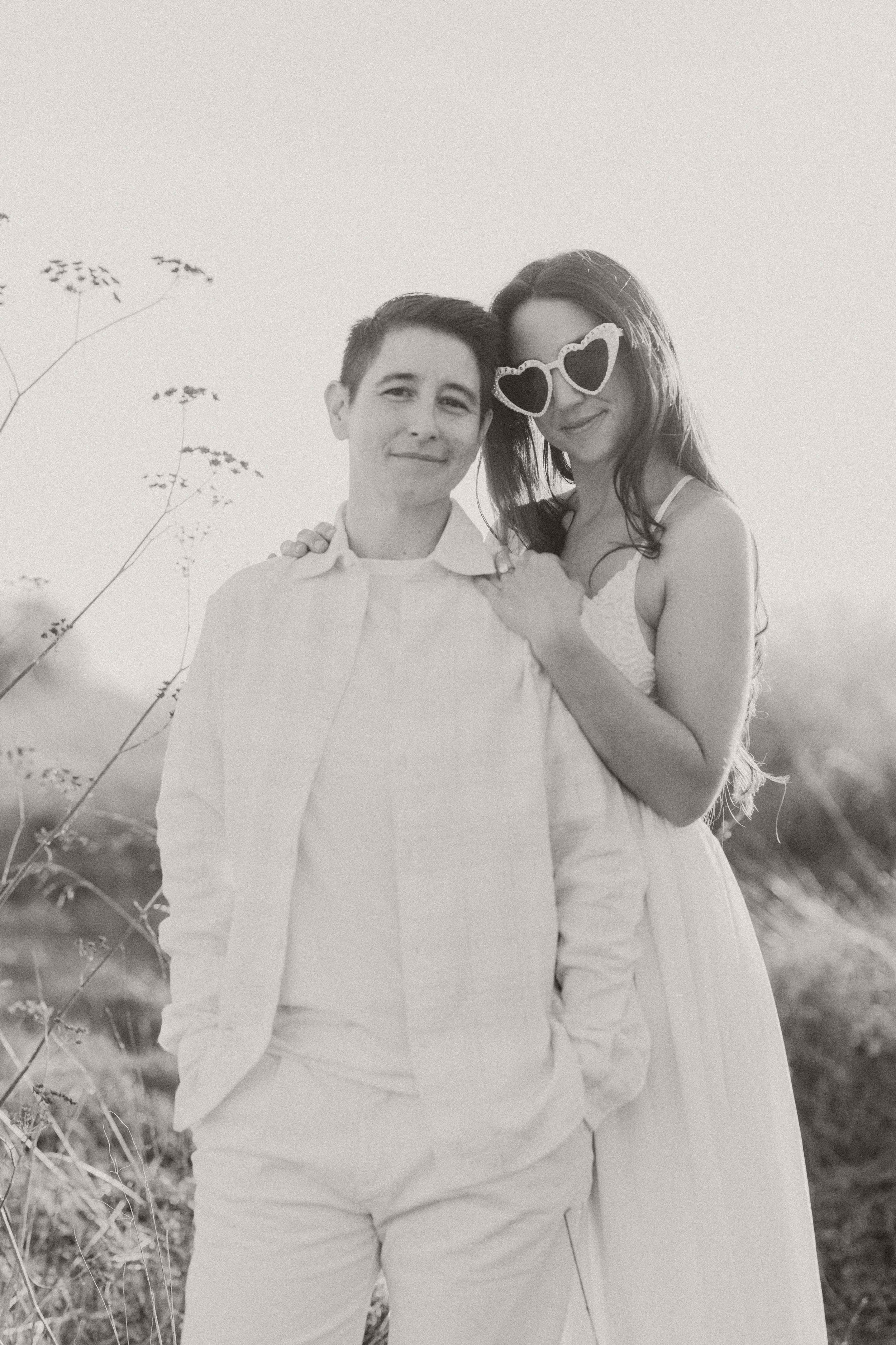 Couple standing close together in a sunlit field, one wearing heart-shaped sunglasses
