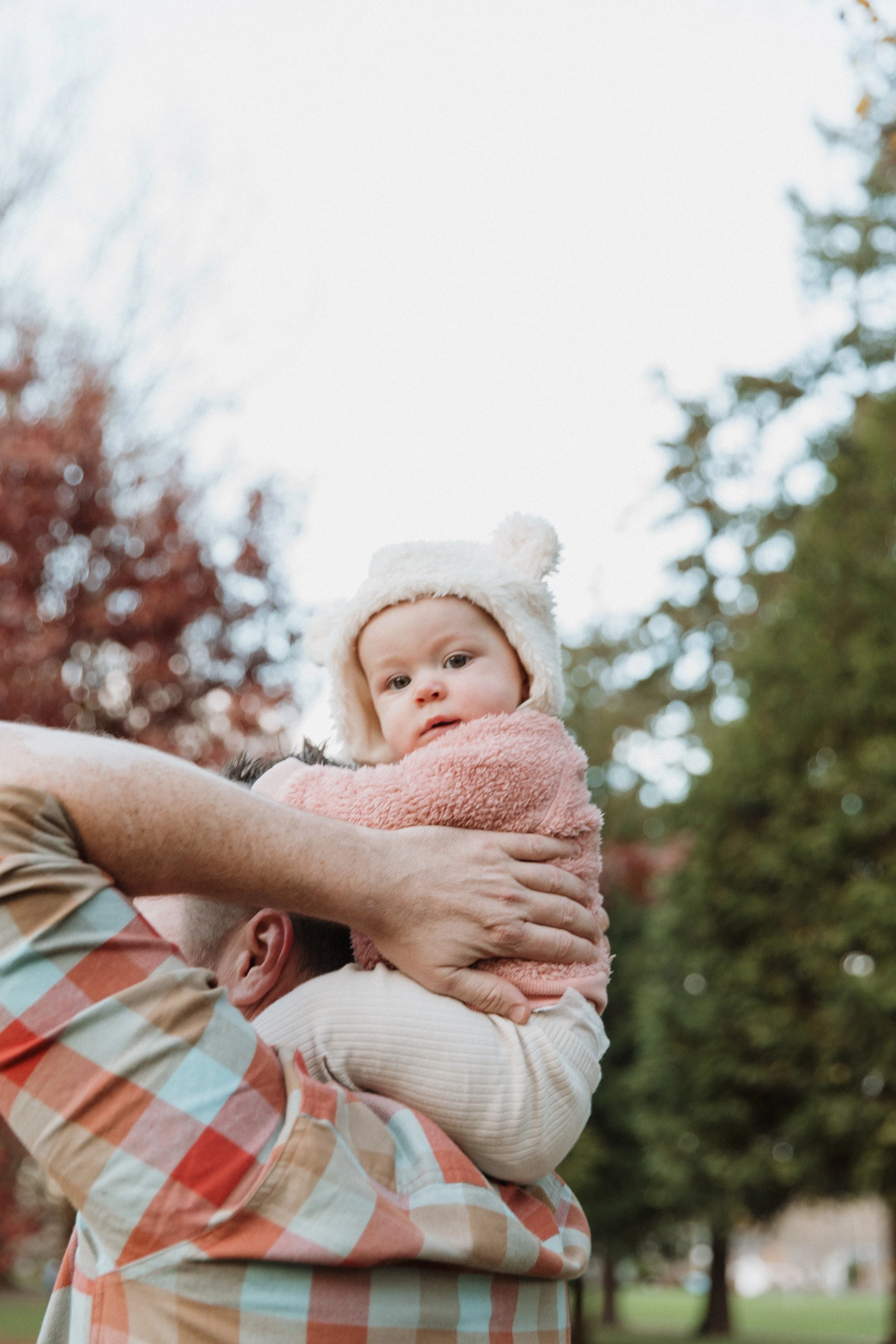 Baby in a bear-ear hat held up by a parent among fall trees